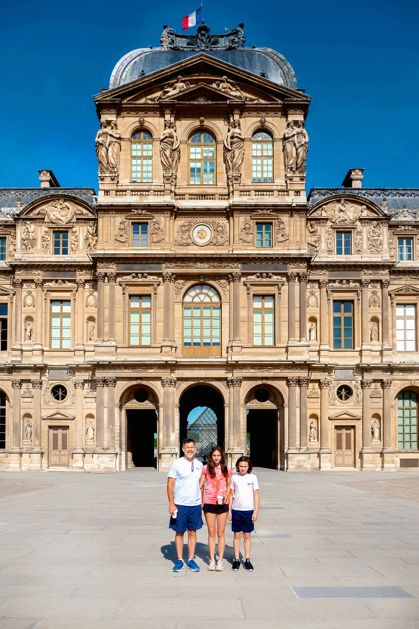 Blog post about things not to forget when traveling to Europe.  The image shows a dad and two kids standing in central Paris, France.  They are standing in front of the back side of the Louvre. 