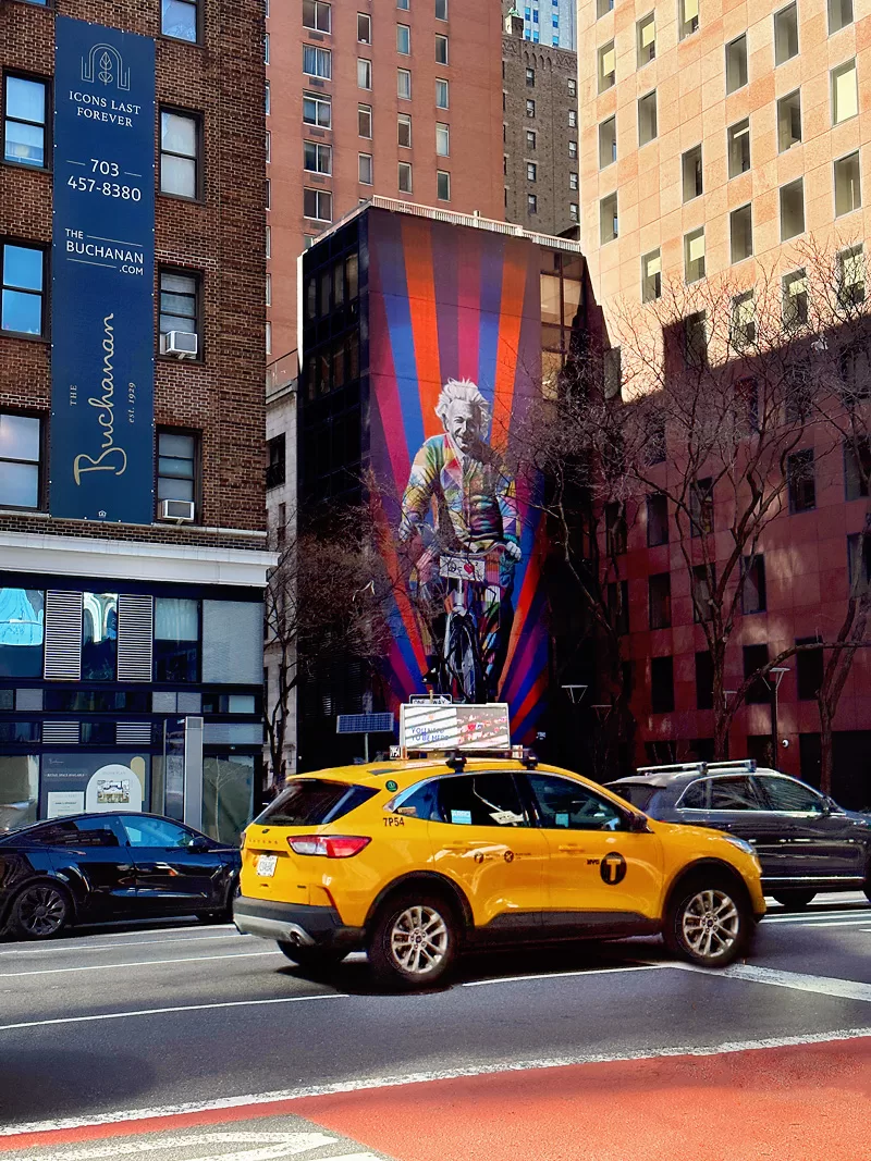 A photo taken in Midtown East NYC. A yellow cab is seen driving past the Einstein mural.