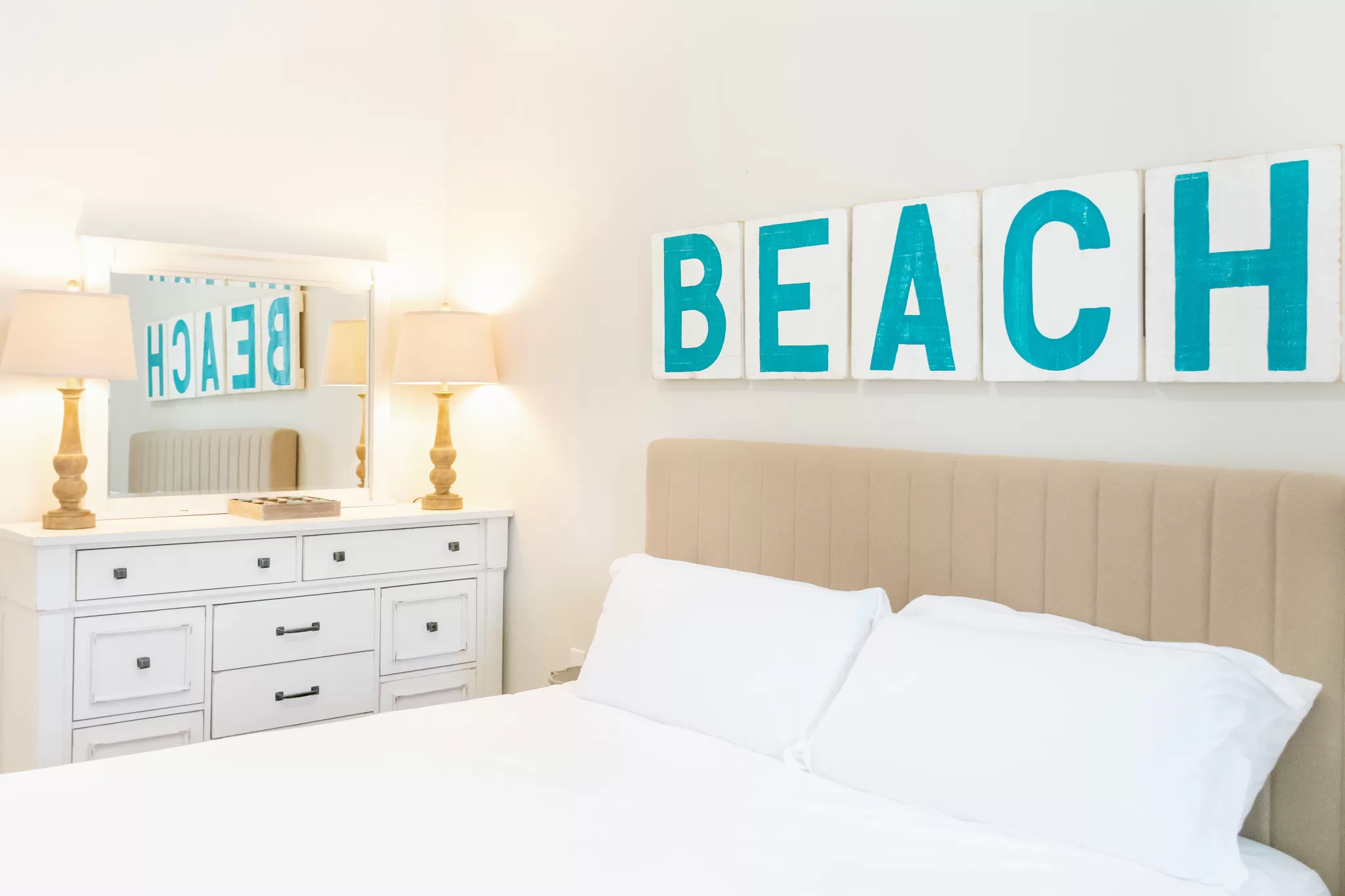 In this photo you see a King size Zinnus bed with white bedding. In the back is a dresser. Letters spelling out the word beach are above the bed.