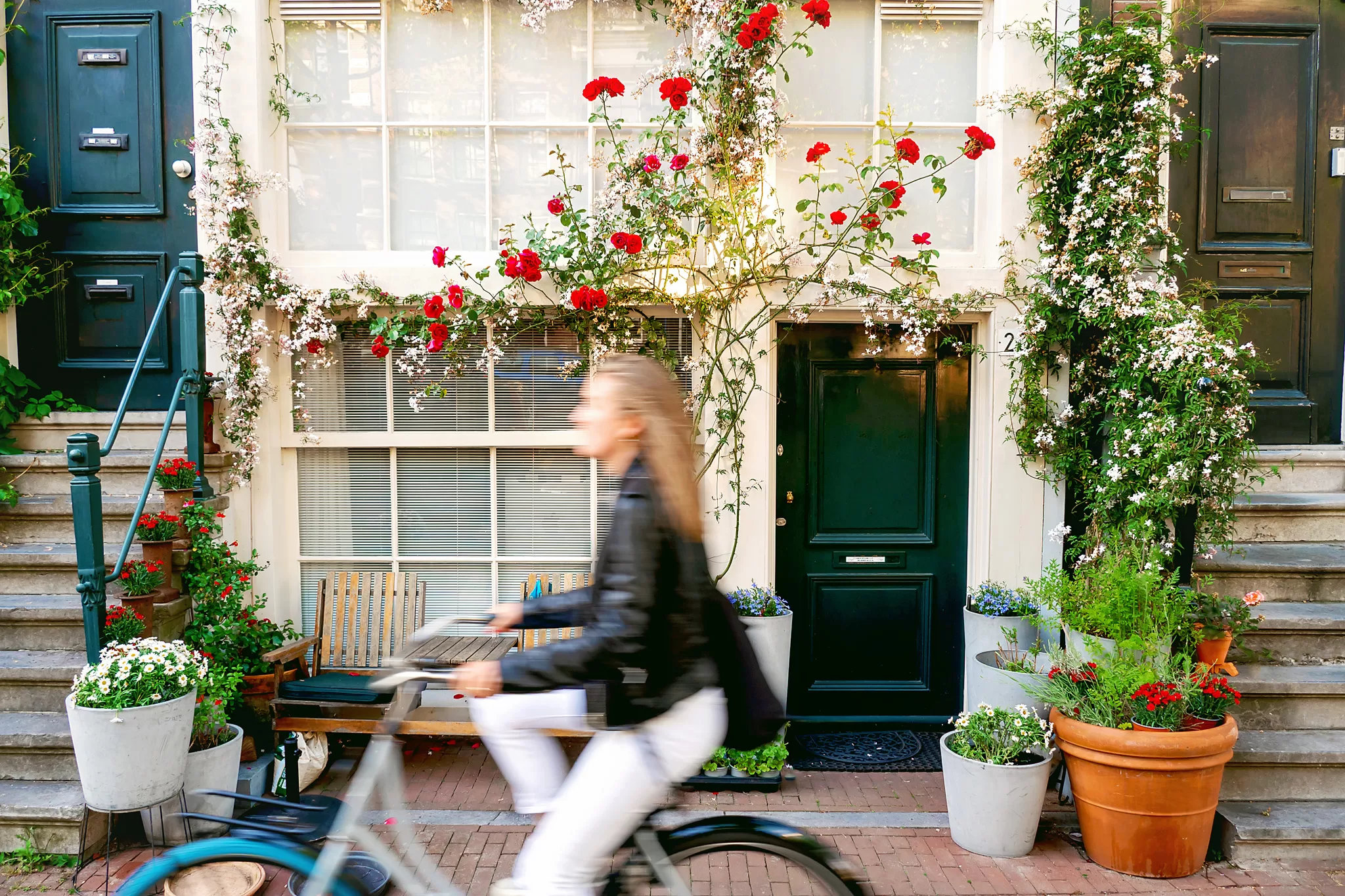 Blog post about travel necessities for Europe Trip.  This image was taken in Amsterdam and shows a building with beautiful flowers on and around it.  A woman was riding by on her bike at the same time the photo was taken and is passing by in a blur. 