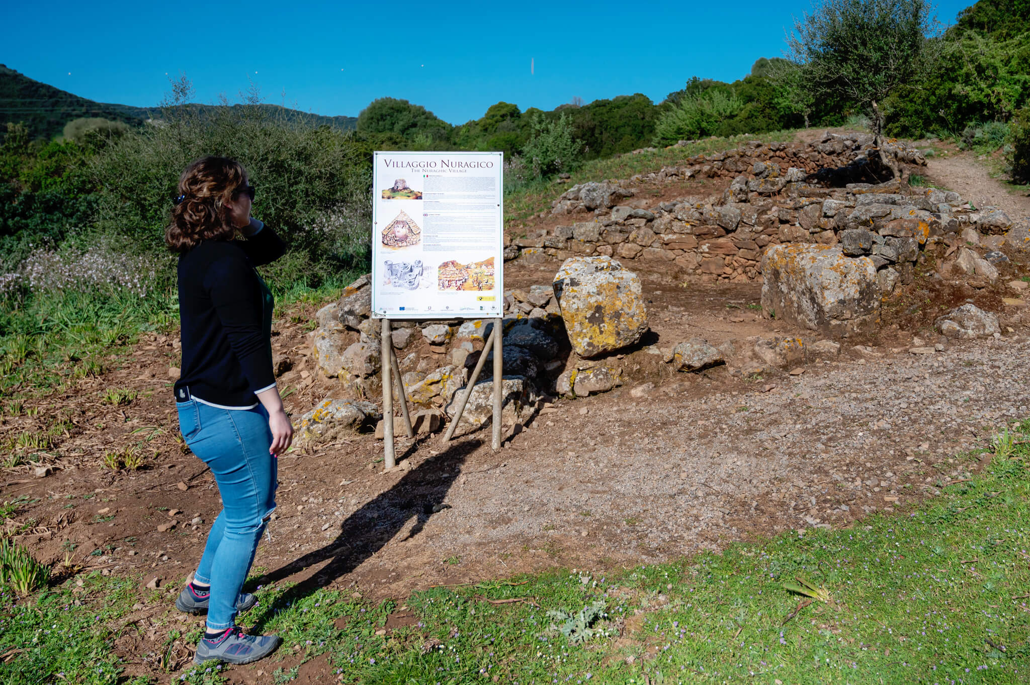 Person in blue jeans and a black jacket stands at left, reading an informational sign beside a short stone ruin on a sunny trail.