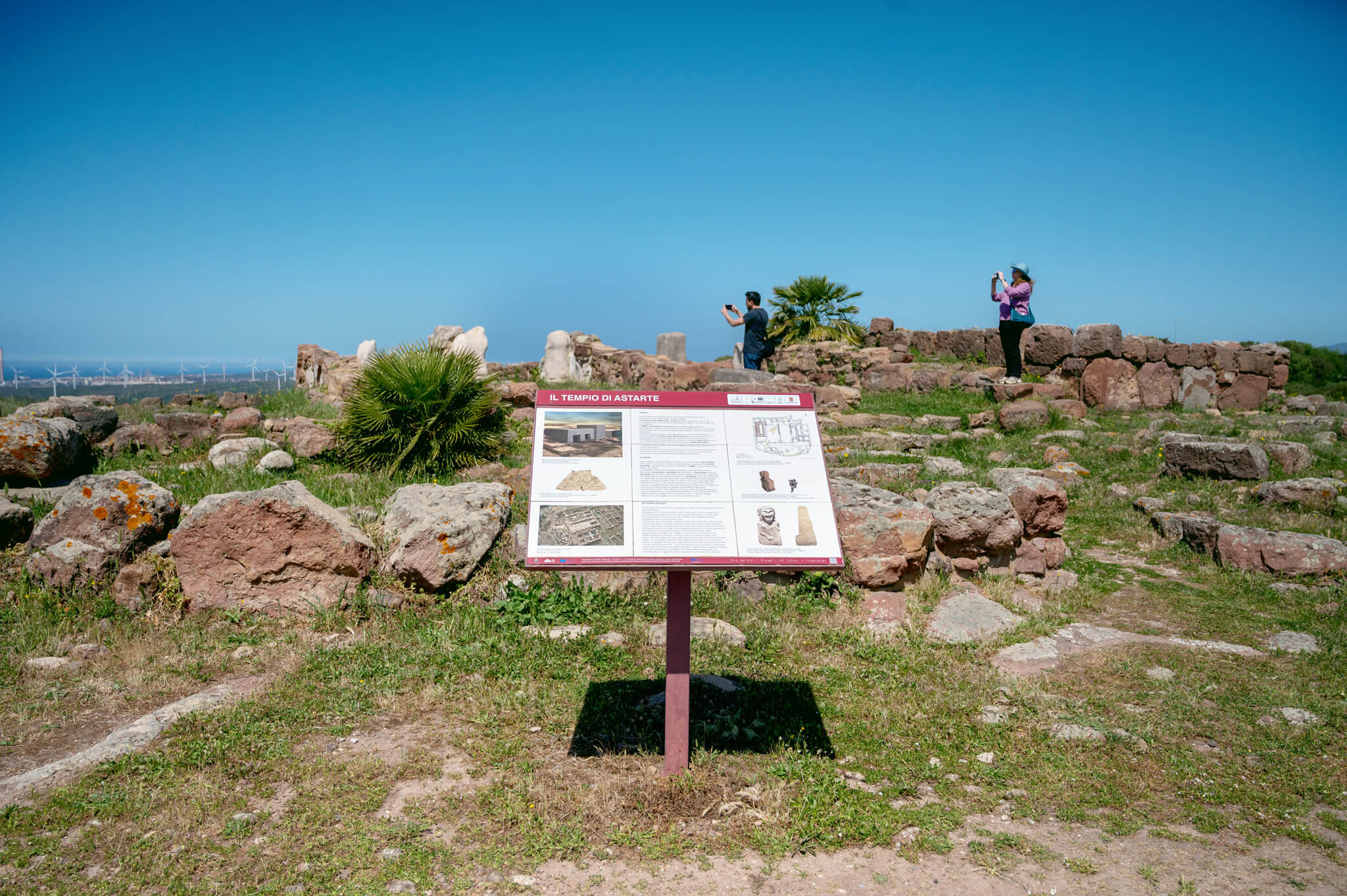 Info board at an archaeological ruin site with two visitors taking photos among scattered rocks under a clear blue sky.