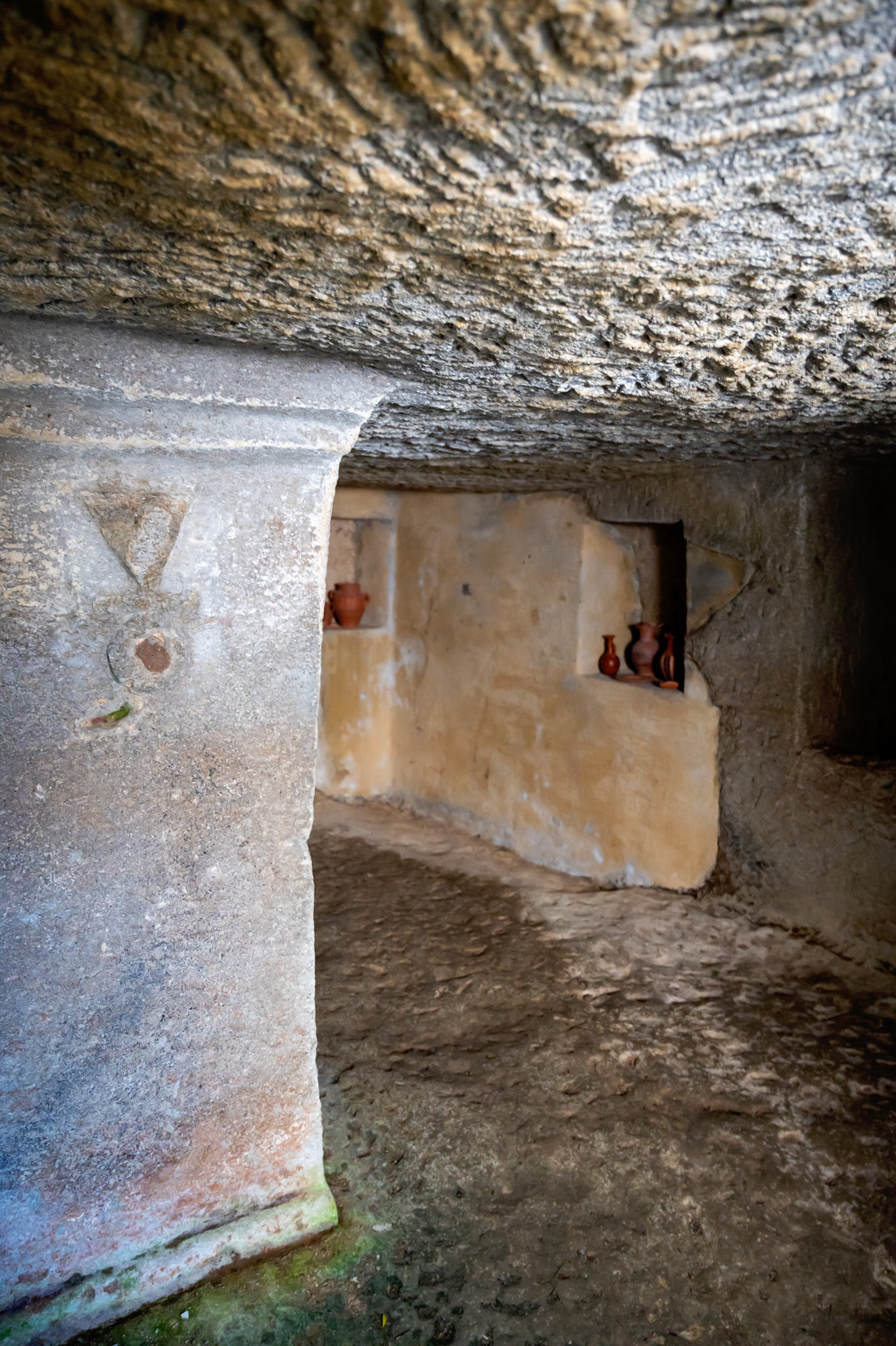 Interior of a rough stone cave with a prominent pillar in the foreground and small clay pots on a recessed shelf in the background.
