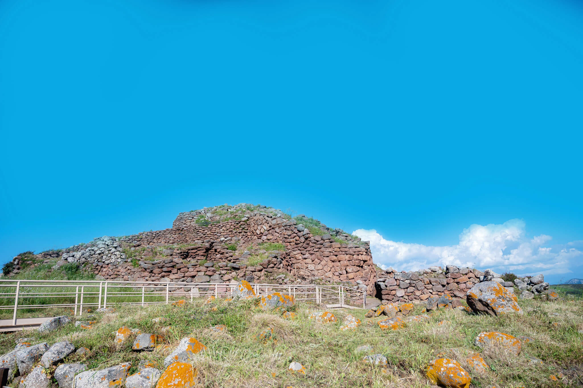 Stone ruin built of layered rocks on a grassy hill, with a white railing in the foreground and a bright blue sky above.