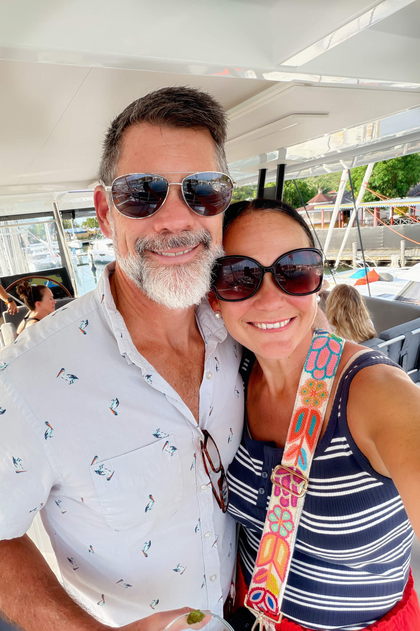 Smiling couple taking a selfie on a boat; man with sunglasses and gray beard, woman with oversized sunglasses and a colorful strap across her chest, marina in background.