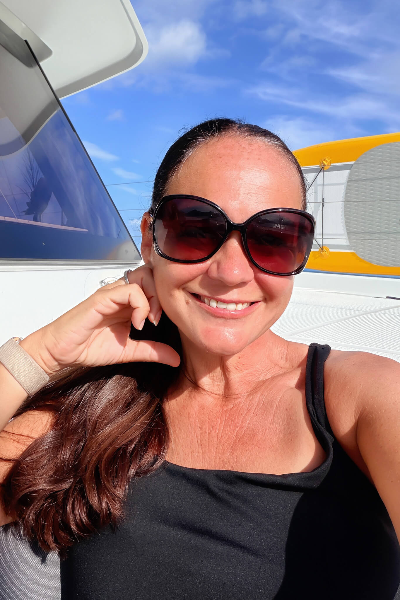 Close-up selfie of a smiling woman wearing large dark sunglasses, with a blue sky and boat deck behind her.