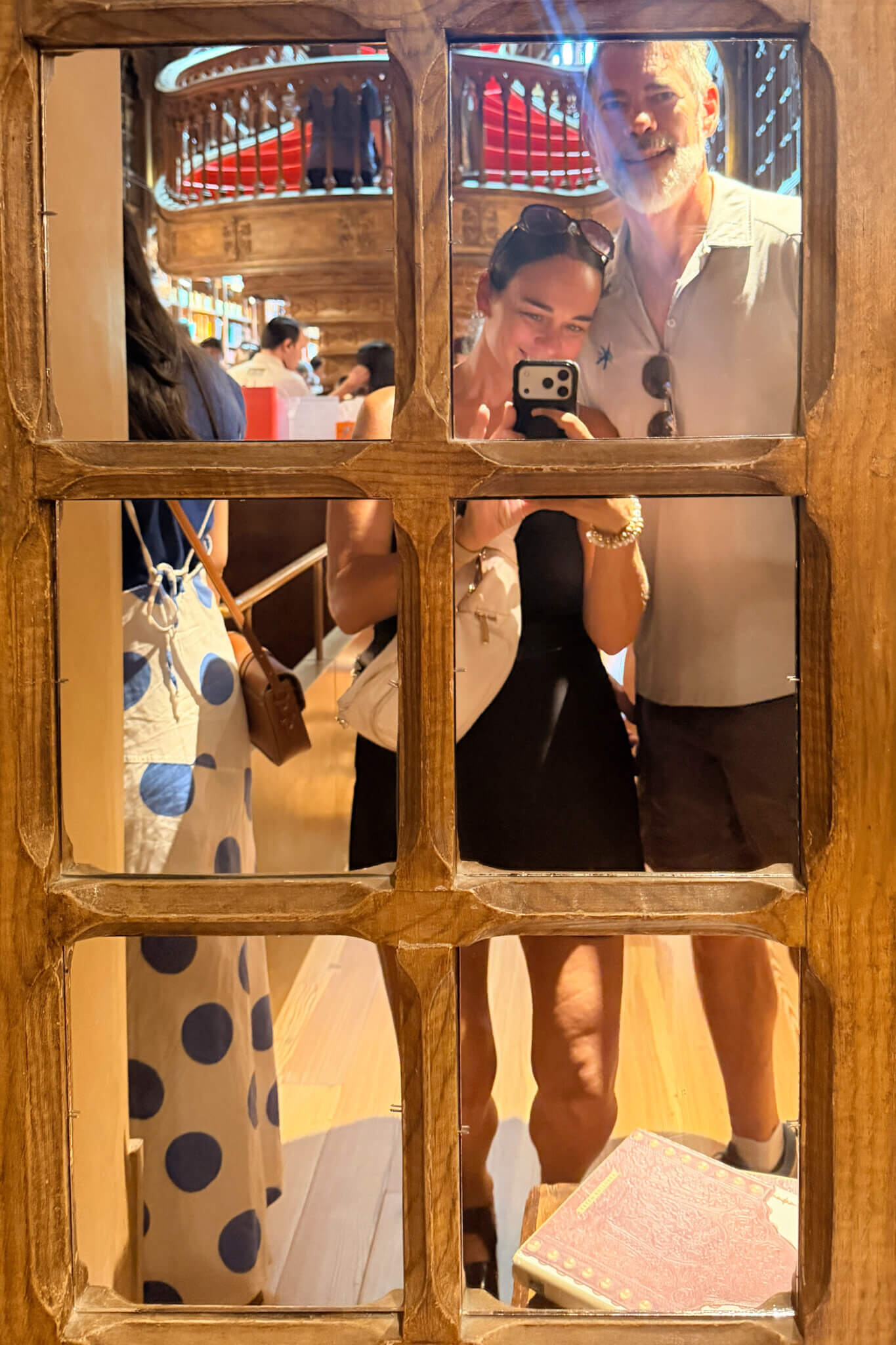 Two people pose for a selfie in a wooden-framed window, smiling with a phone, inside a busy store or cafe with shelves in the background.