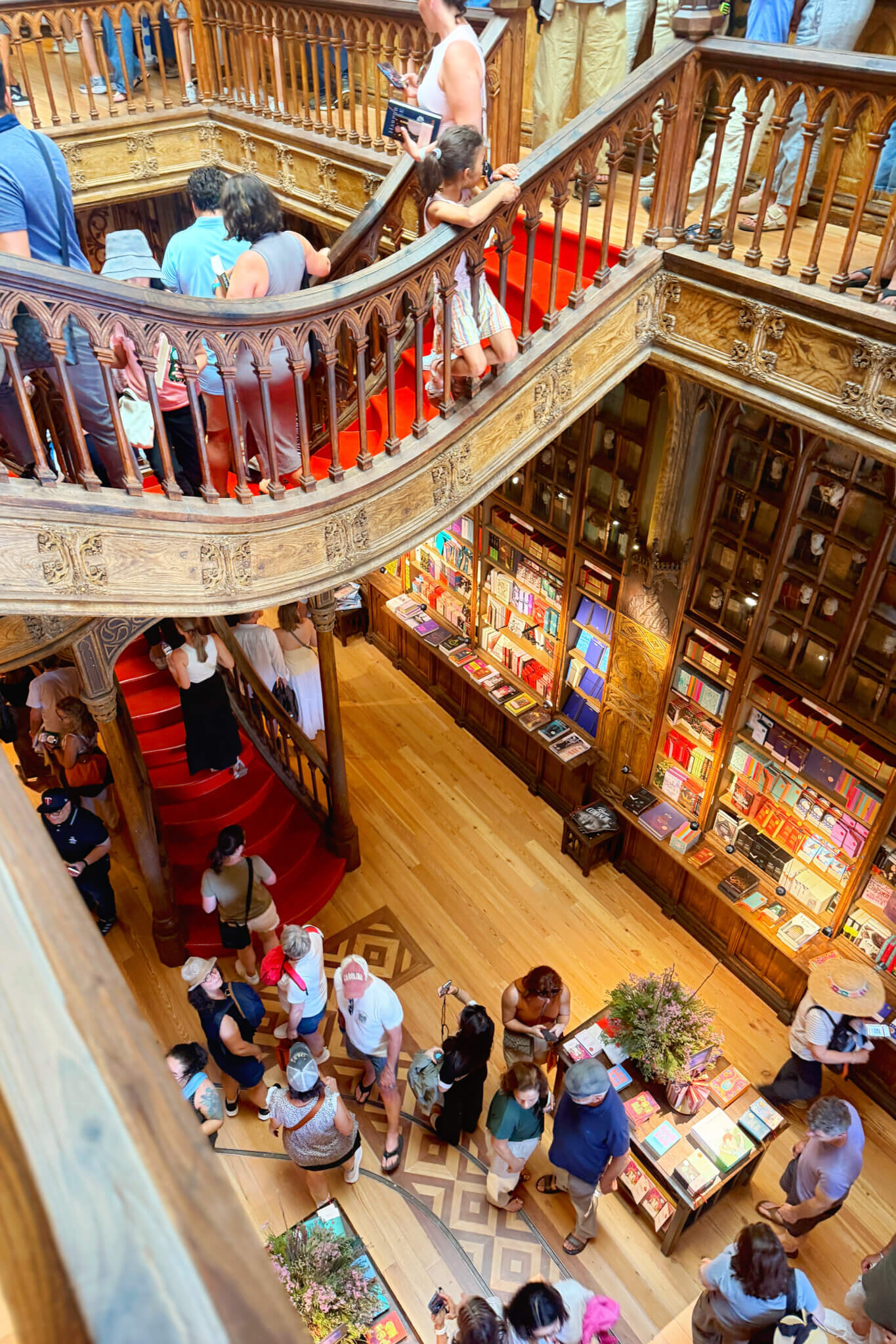 Busy multi-level bookstore with an ornate wooden spiral staircase and red carpet, people browsing shelves full of books on both levels.