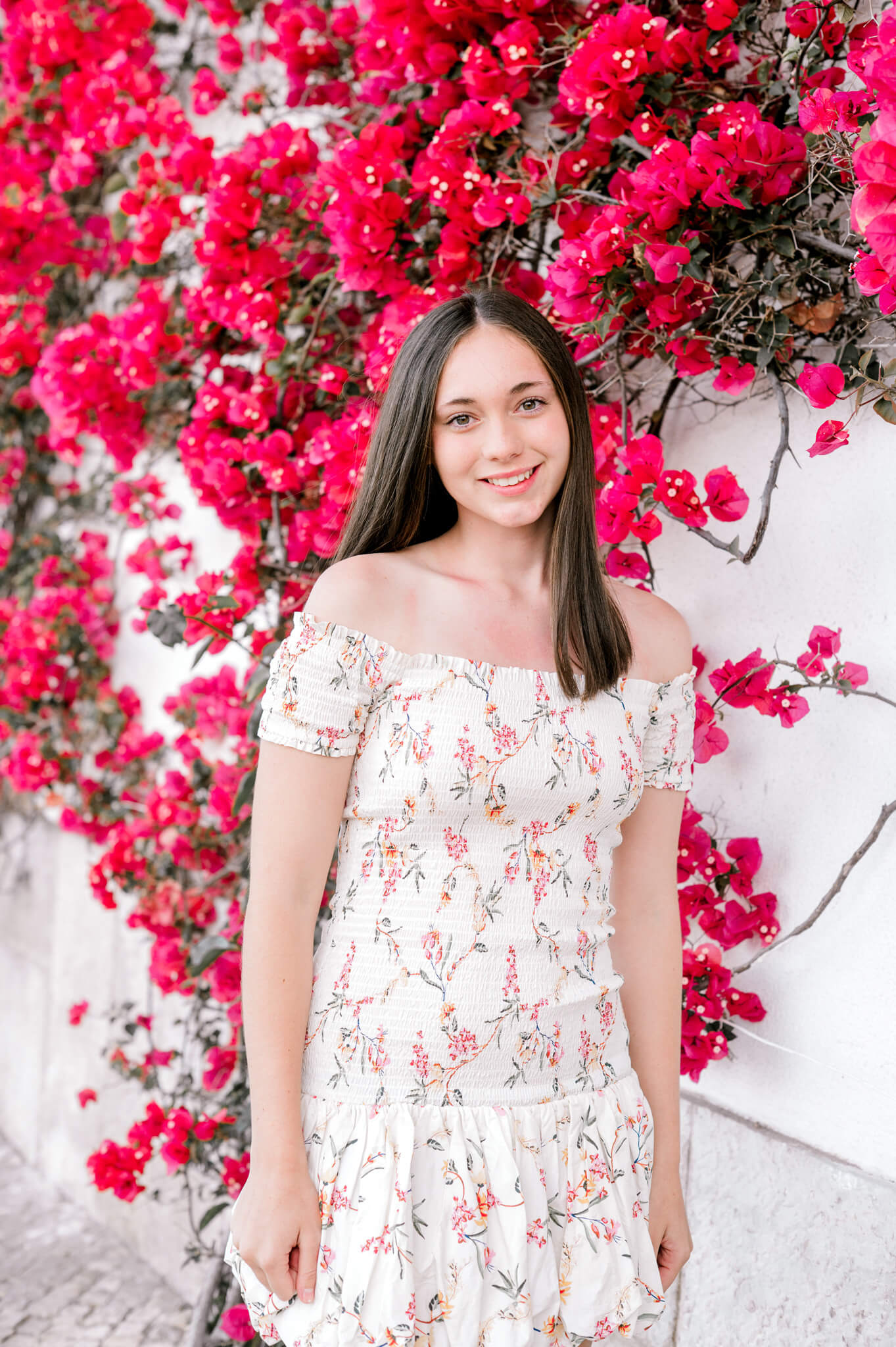 Young woman in a white floral off-shoulder dress stands smiling in front of bright pink bougainvillea vines.