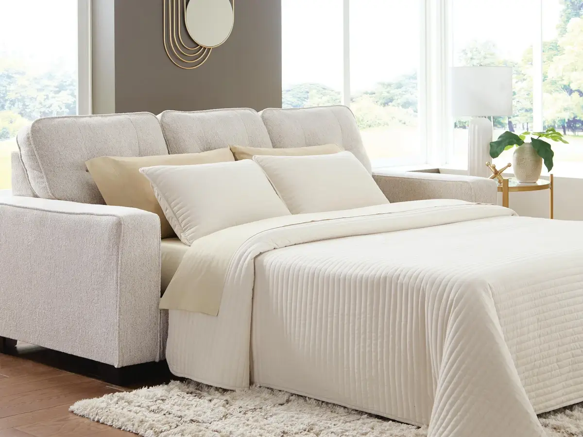 Cream bed with a white quilted coverlet and beige pillows beside a gray upholstered sofa in a bright, sunlit bedroom.
