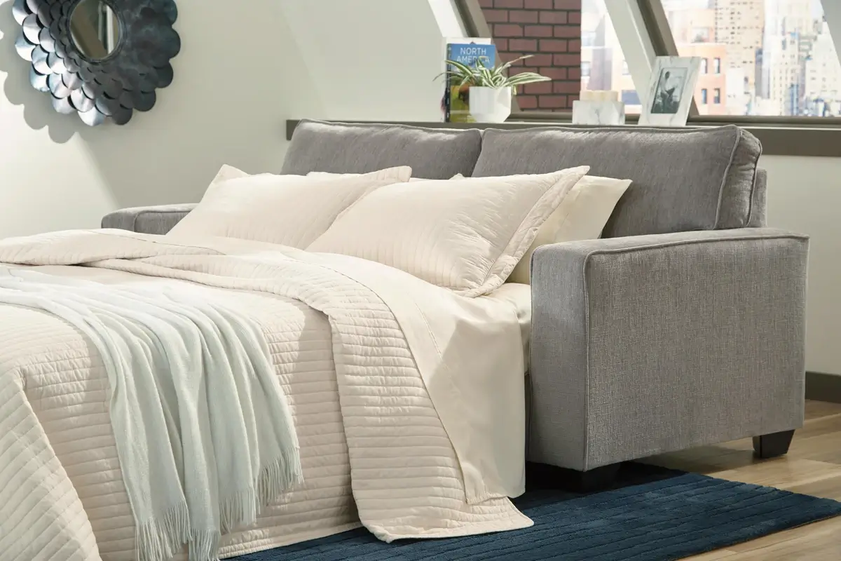 Grey upholstered bed with white bedding and a pale blue throw on a dark blue rug in a bright urban bedroom with a city view.