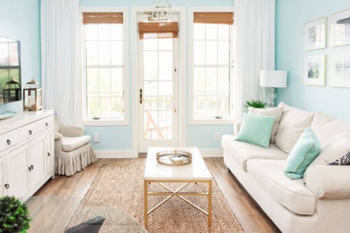 Bright living room with light blue walls, white sofa and mint cushions, glass coffee table, and large windows with bamboo shades at the back.