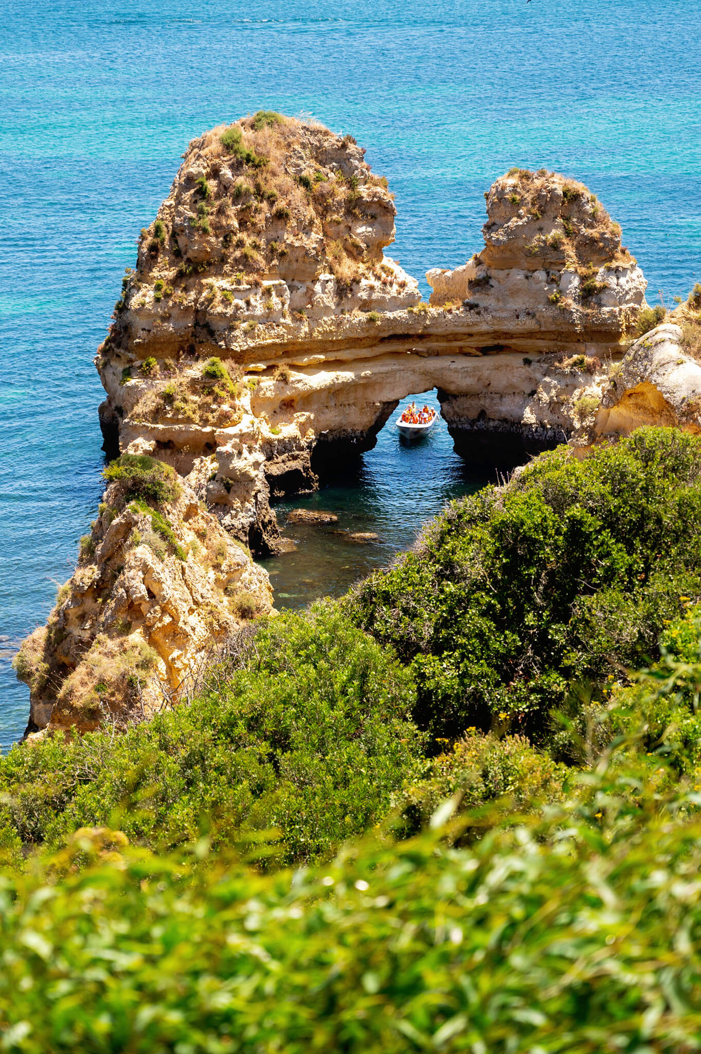Rock arch along the turquoise sea with a small boat passing beneath, edged by green shrubs in the foreground