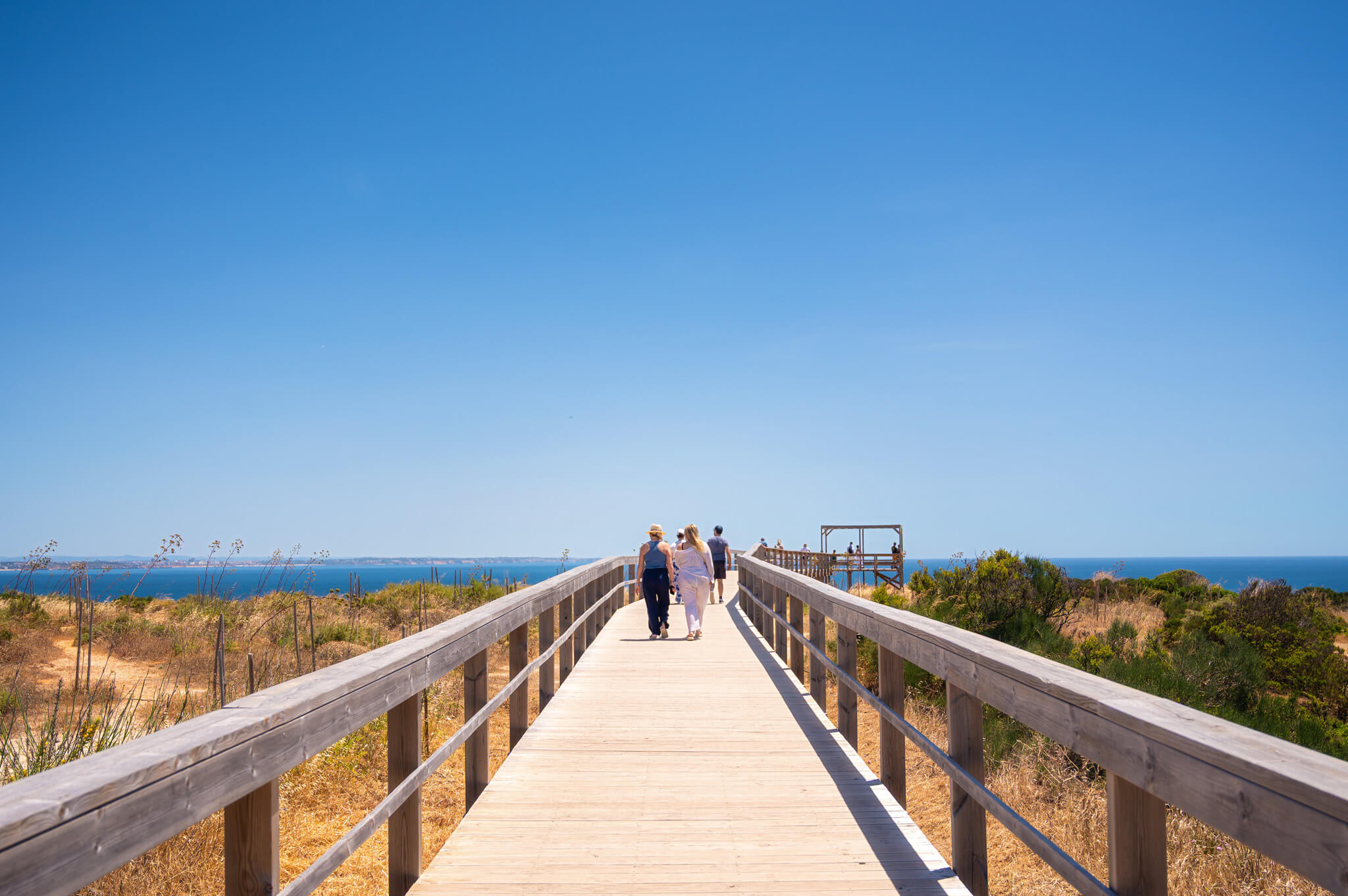 Group of people walking along a long wooden boardwalk toward the blue ocean under a clear sky, with dry grasses on either side.