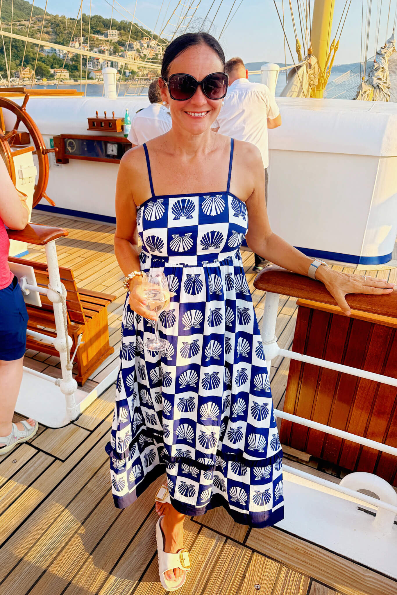 Woman in blue-and-white patterned sundress and sunglasses on a yacht deck, holding a wine glass with a harbor in the background.