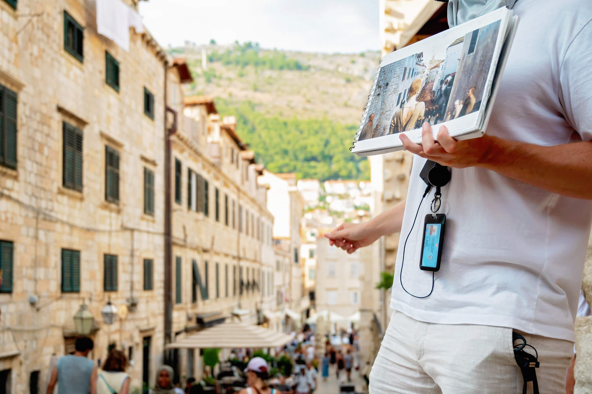 Tour guide holding a photo album in a sunlit historic stone street, wearing a lanyard and badge.