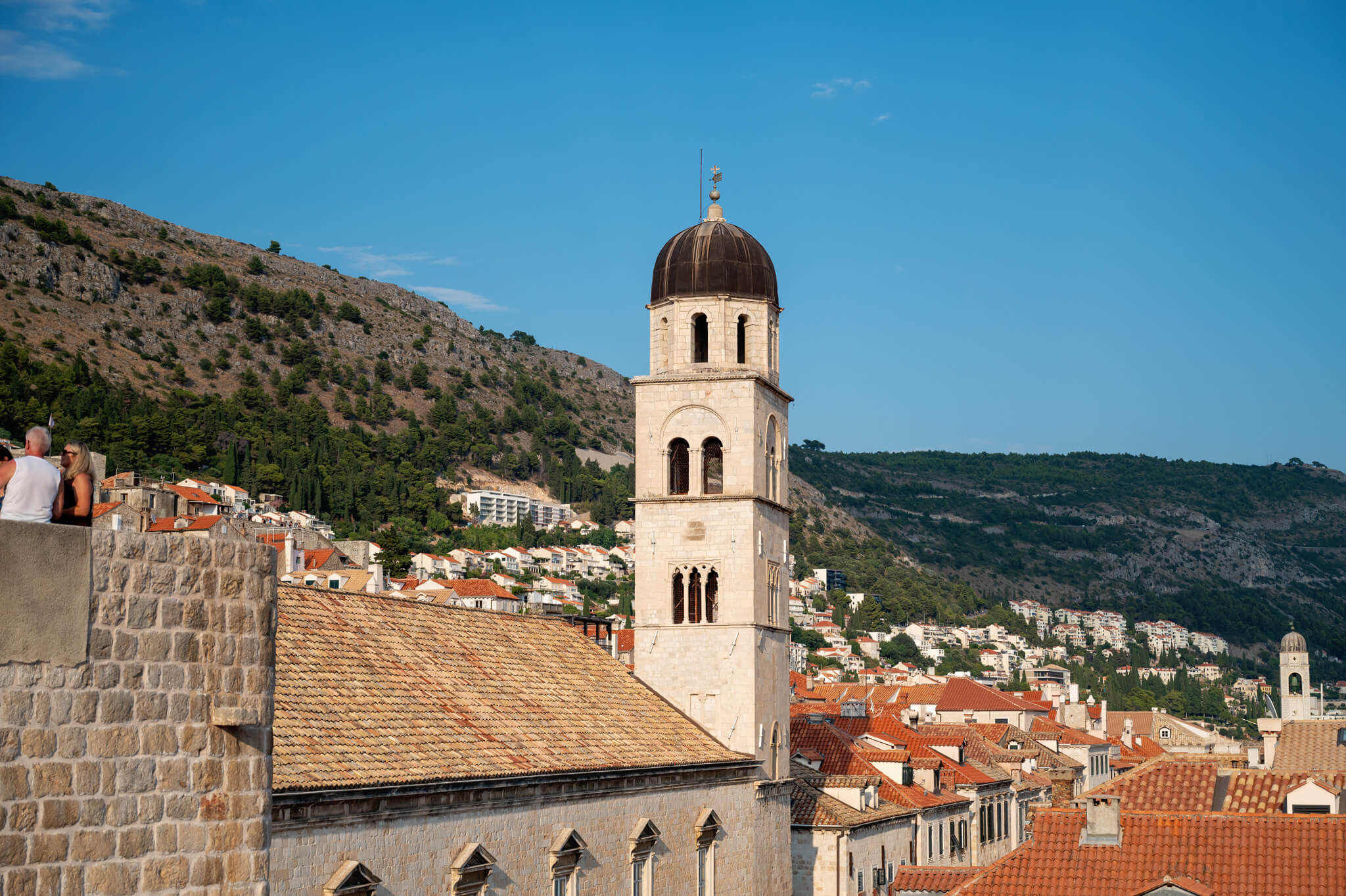 Stone bell tower with a dark dome rises over a town of red-tiled roofs against a clear blue sky, hillside in the background, people seen at left edge.
