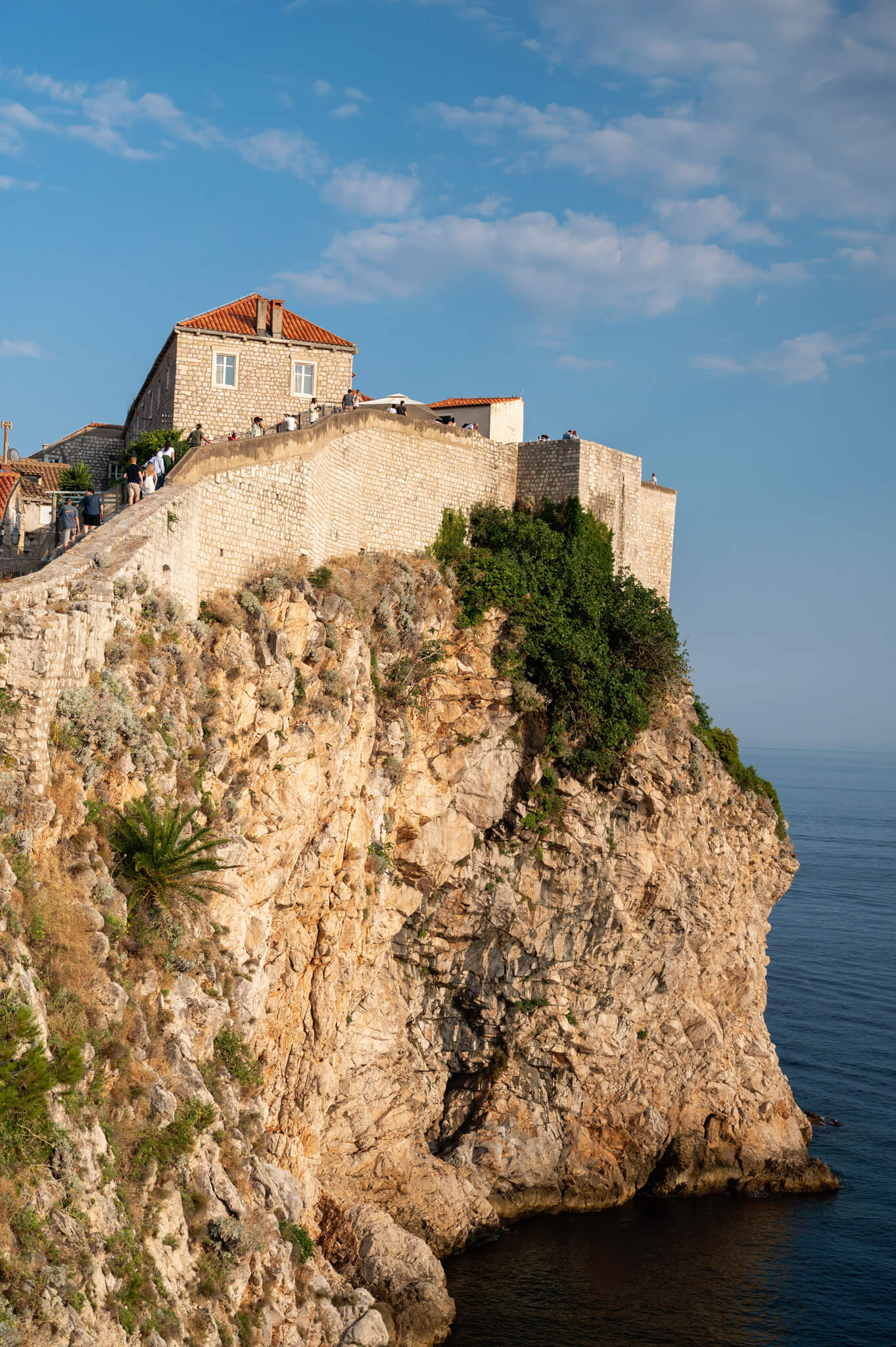 Stone fortress and curved wall on a cliff, with visitors walking along the rampart under a blue sky.