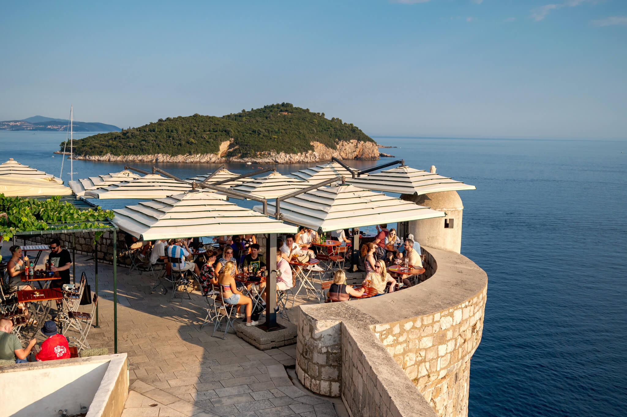 Outdoor seaside café with many people seated at tables under stacked white umbrellas on a stone terrace overlooking calm blue sea and a green rocky island in the distance.
