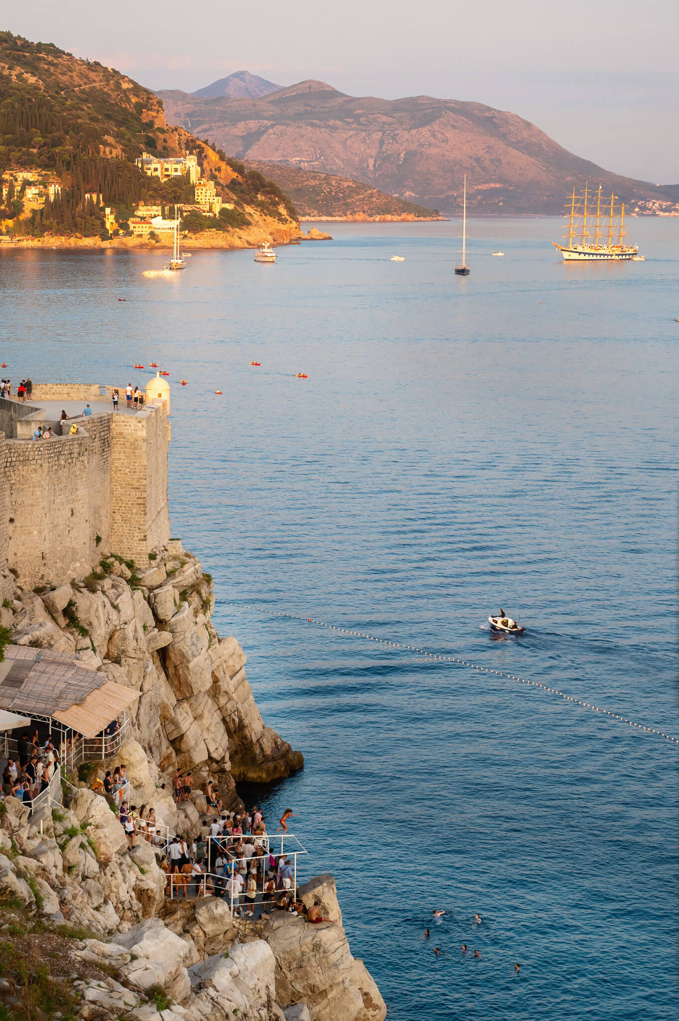 Crowded rocky cliff promenade with people overlooking the calm blue sea and distant mountains.