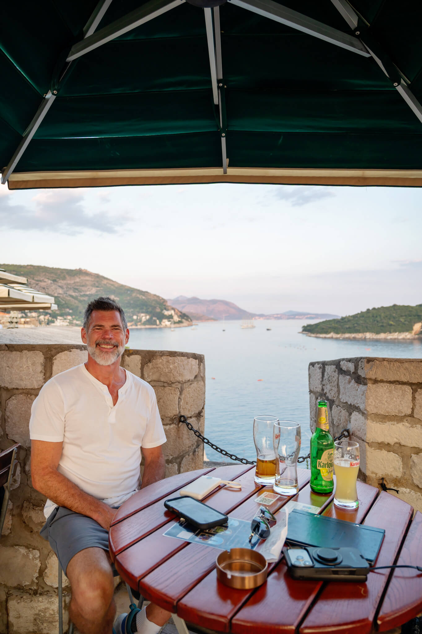 An older man in a white polo sits at a wooden outdoor table by the water, smiling as drinks and glasses sit on the table with a bottle of beer nearby.
