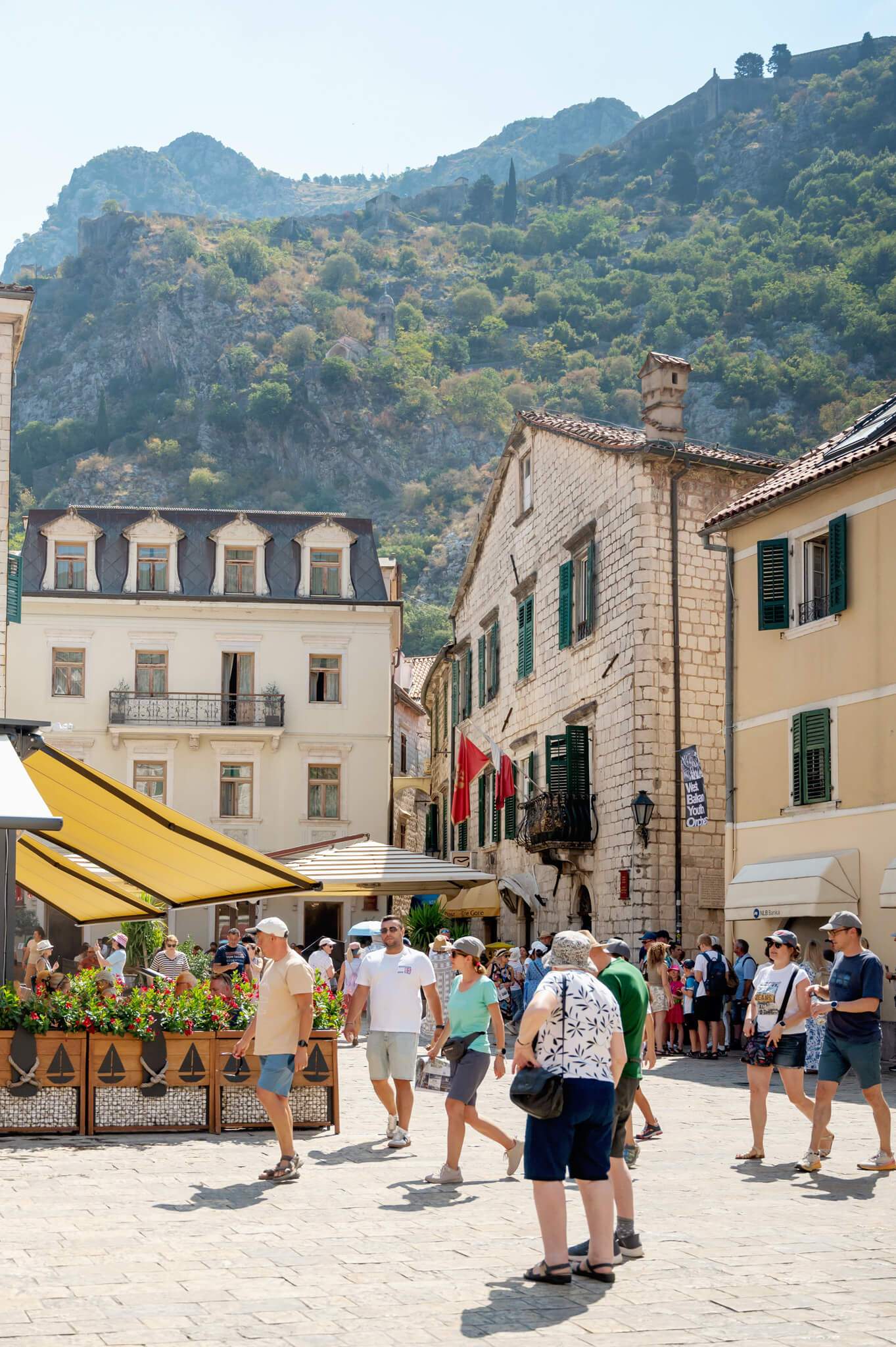 Busy European street scene in Kotor Montenegro in a sunny town square with people walking past cafés and stone buildings with green shutters, mountains in the background.