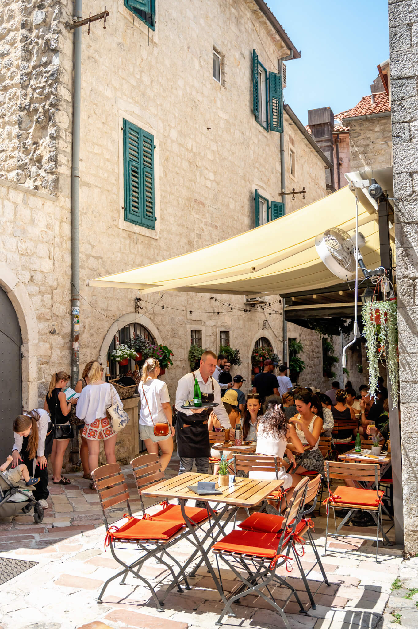 Kotor Montenegro Outdoor stone-walled alley cafe with green shutters; people sit at wooden tables under a pale yellow canopy enjoying drinks and meals.