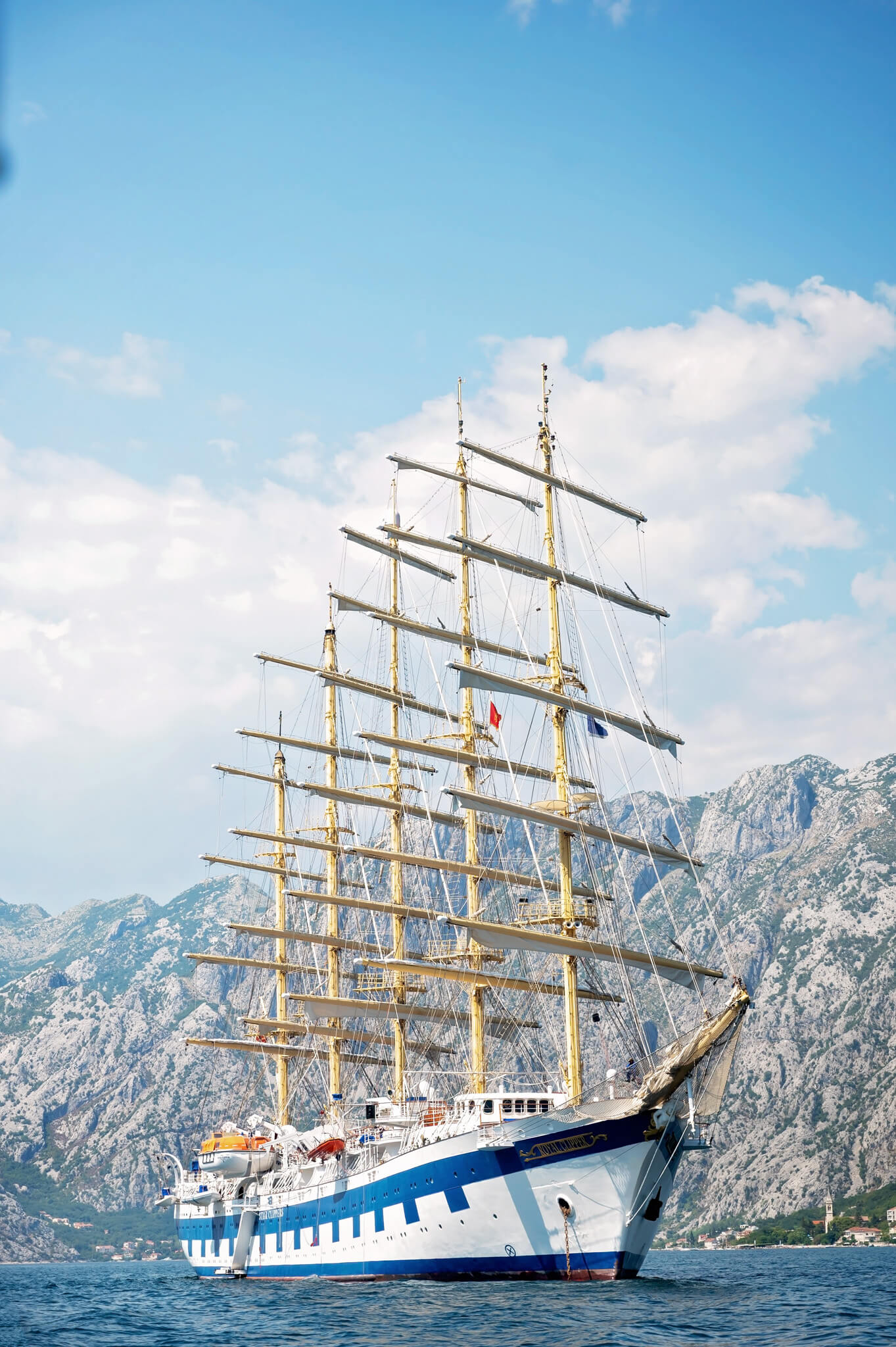 Star Clippers Royal Clipper Day 4 Kotor Montenegro 10 • Our Little Lifestyle A large Royal Clipper multi-masted sailing ship moored near a rocky coastline under a blue sky with clouds, its tall masts and rigging towering above the water.
