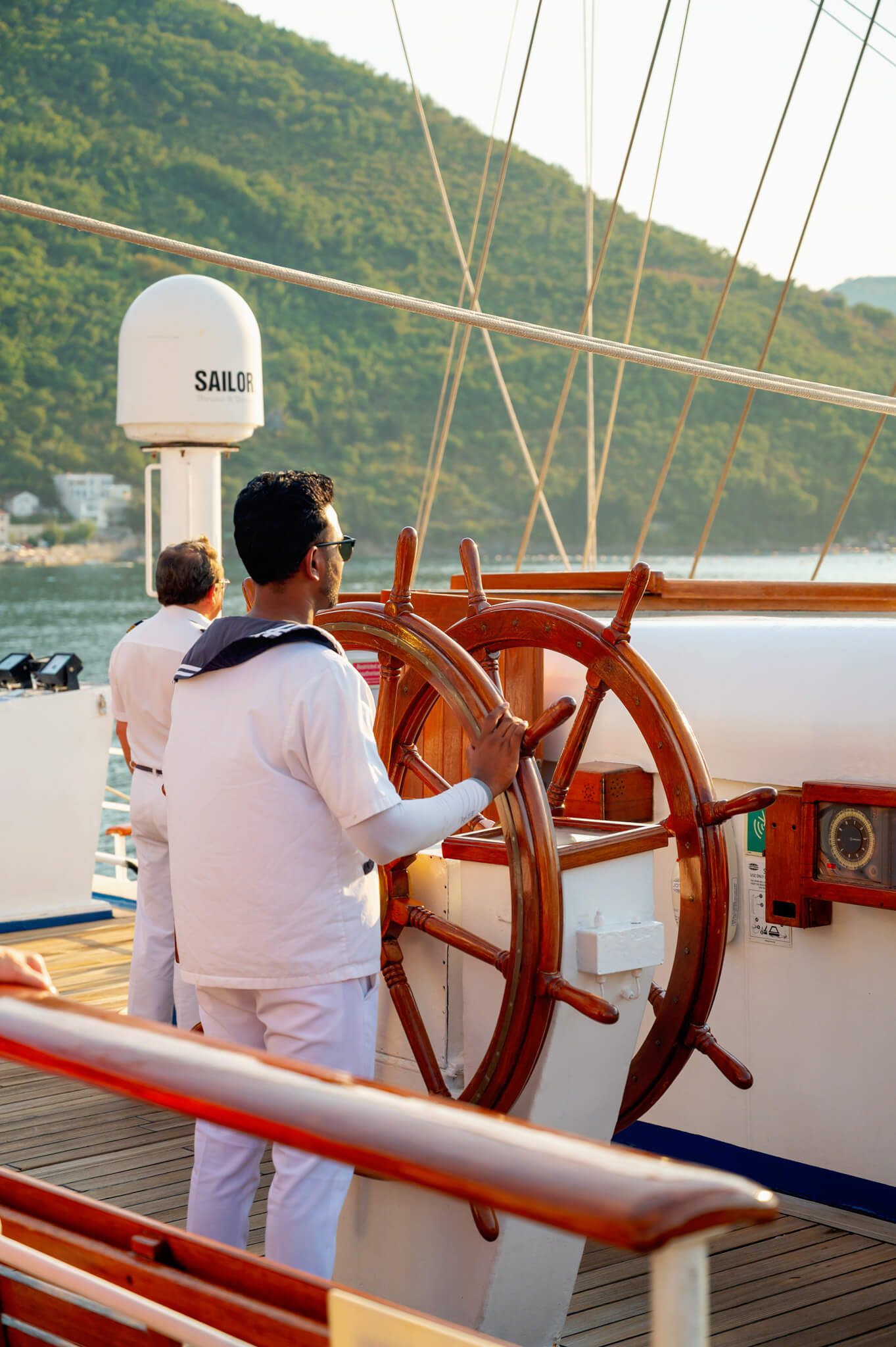 Crew members aboard a sailboat; a man in white steering a wooden helm with another crew member in the background.