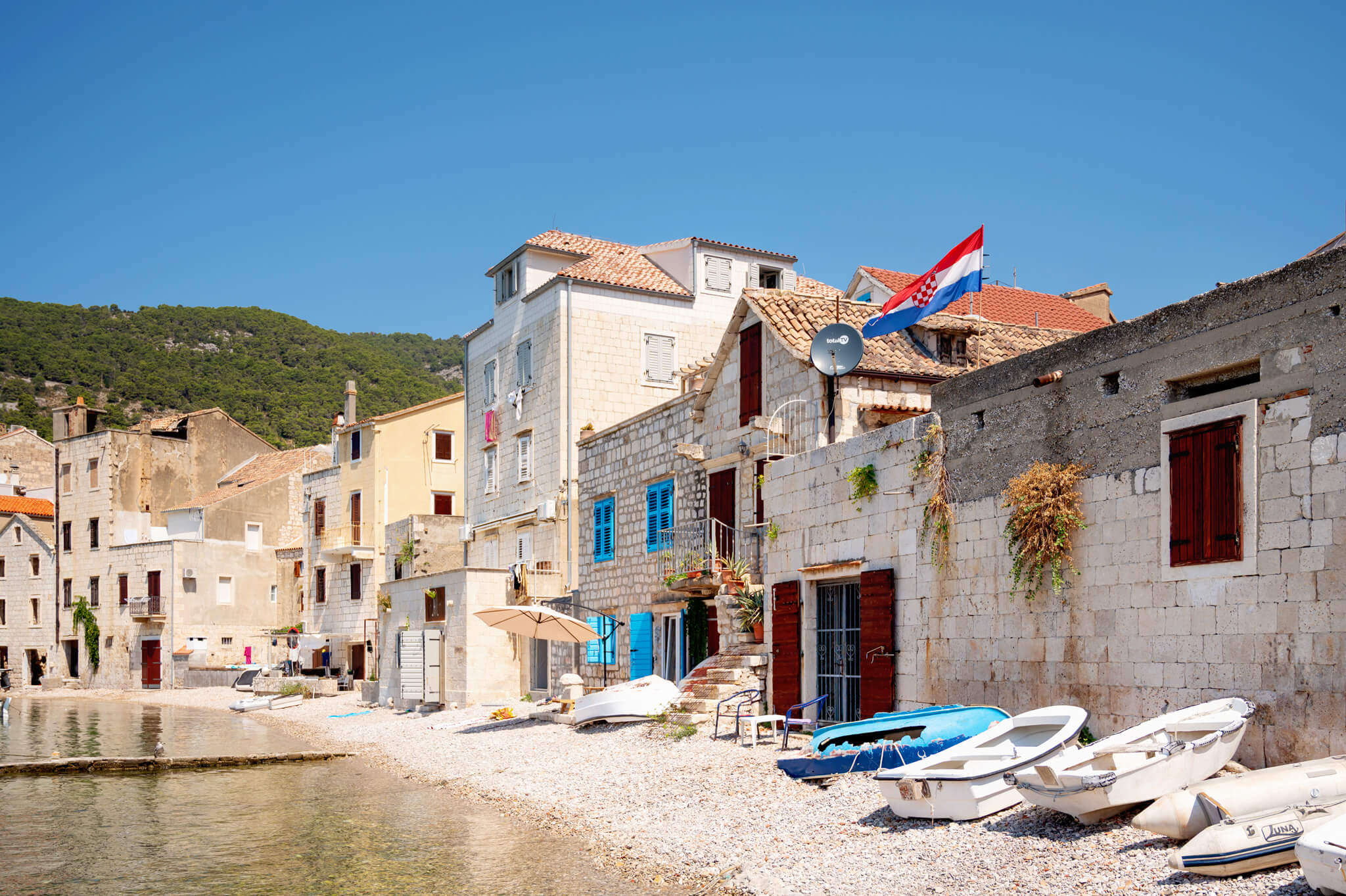 Royal Clipper stop in Komiza, Croatia. Sunny coastal scene with stone houses along a pebble beach and a Croatian flag flying on a house roof.