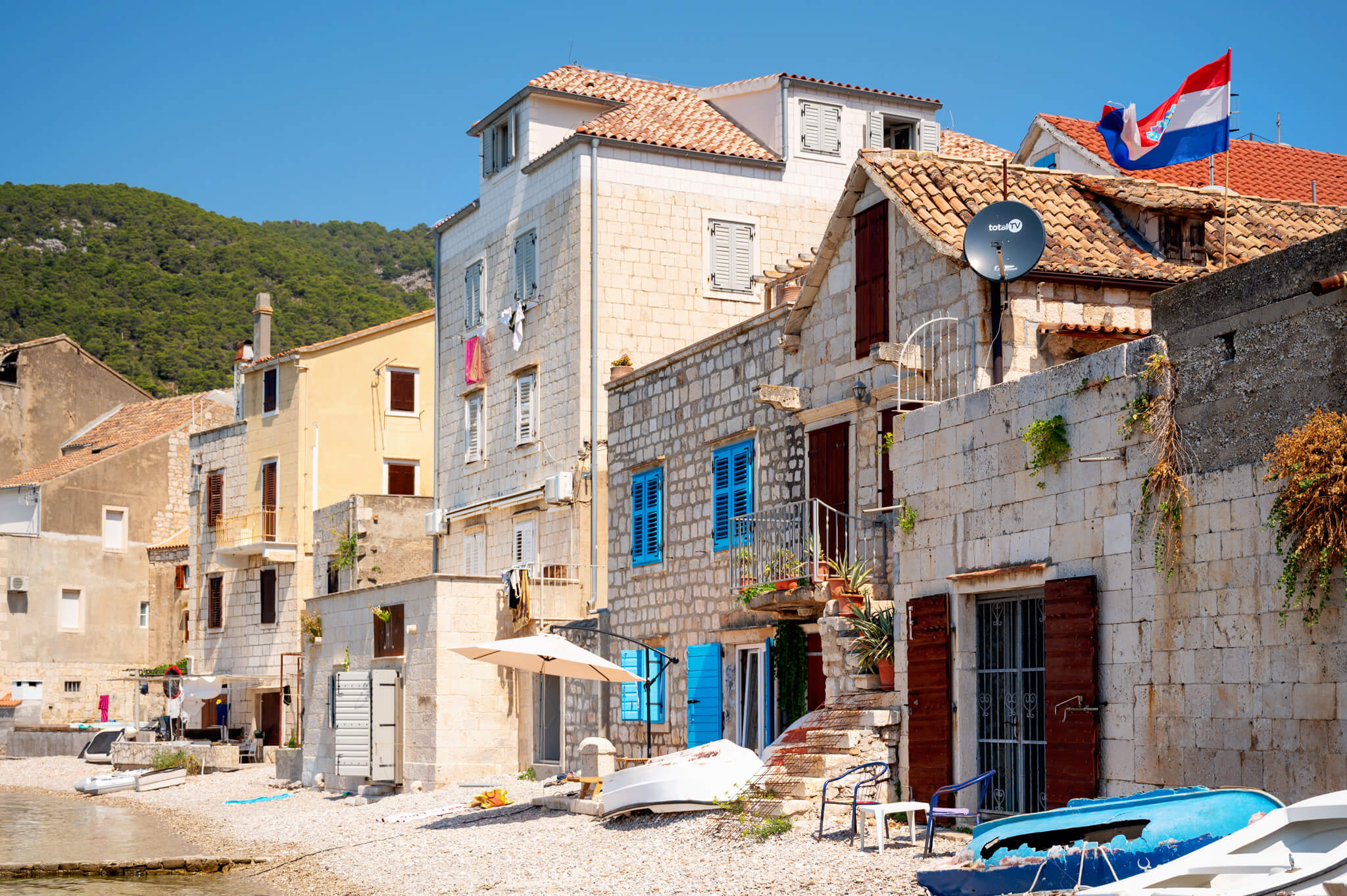 Star Clippers Royal Clipper Day 5 Komiza Croatia 06 • Our Little Lifestyle Royal Clipper stop in Komiza, Croatia. Sunlit seaside street of stone houses with red-tiled roofs and blue window shutters along a pebbled beach under a clear blue sky.