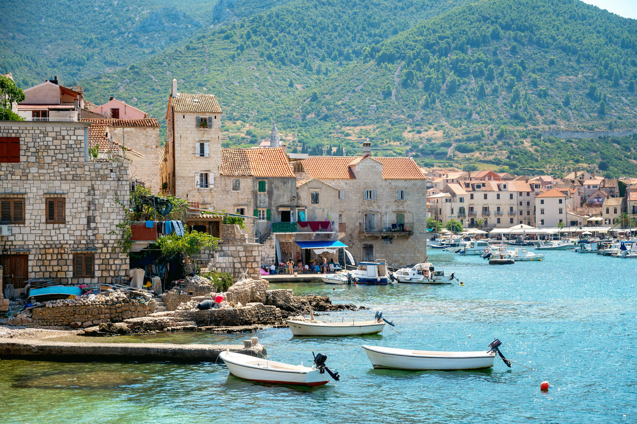 Royal Clipper stop in Komiza, Croatia. Coastal town with stone buildings along a calm harbor and small boats anchored at the quay, gentle hills in the background.