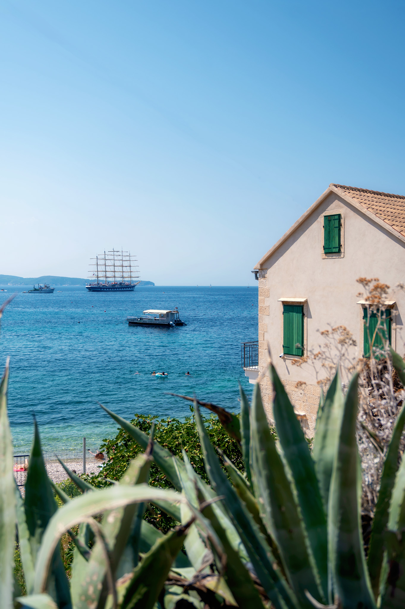 Sea view with a beige house and green shutters on the right, blue sea and a tall sailing ship in the distance; agave plants in the foreground. Royal Clipper stop in Komiza, Croatia.