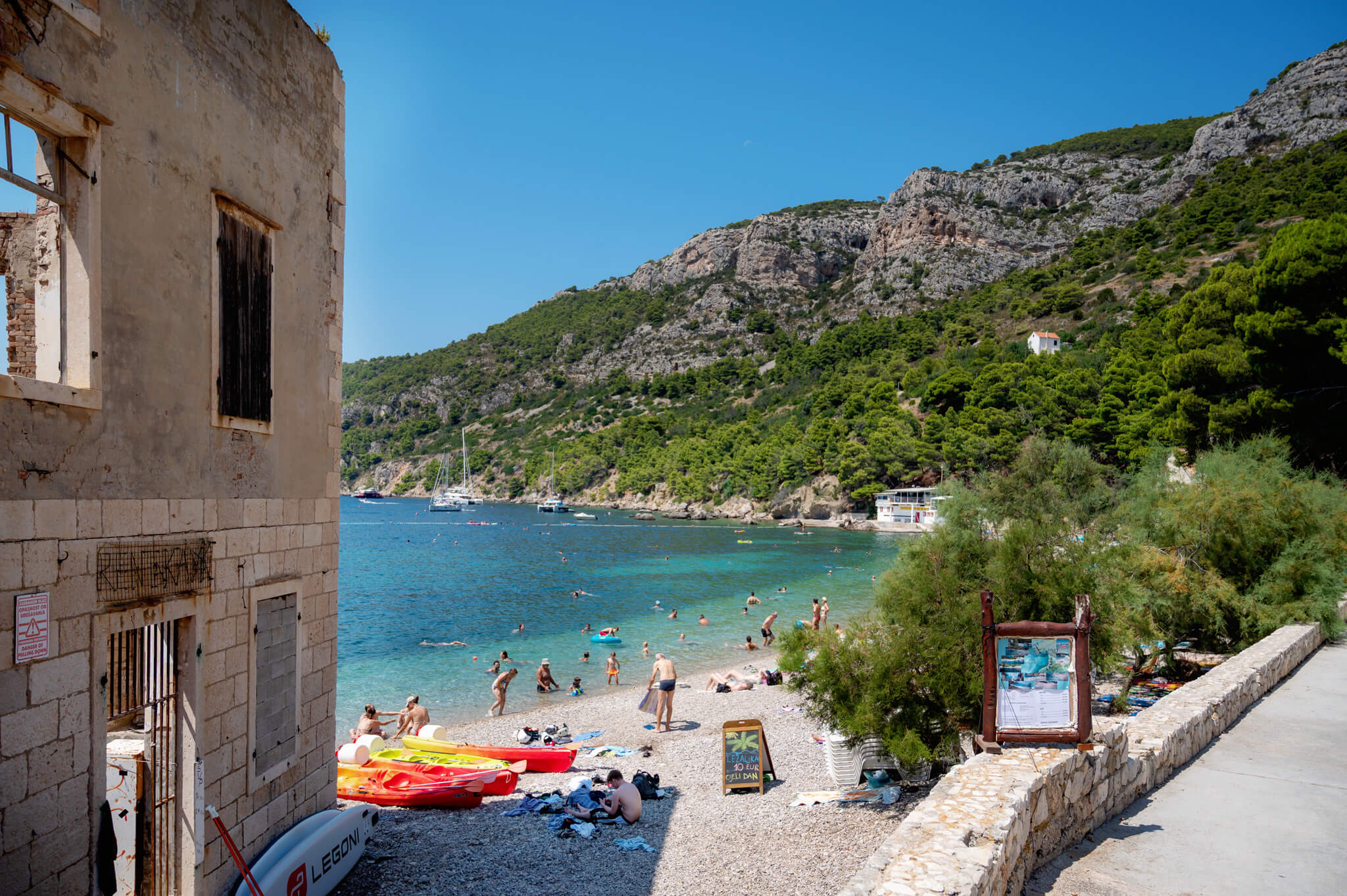 Royal Clipper stop in Komiza, Croatia. Old stone building with a boarded window on a pebble beach, turquoise water and people swimming behind it. A row of colorful kayaks rests on the shore; rocky green hills rise in the background under a clear blue sky.