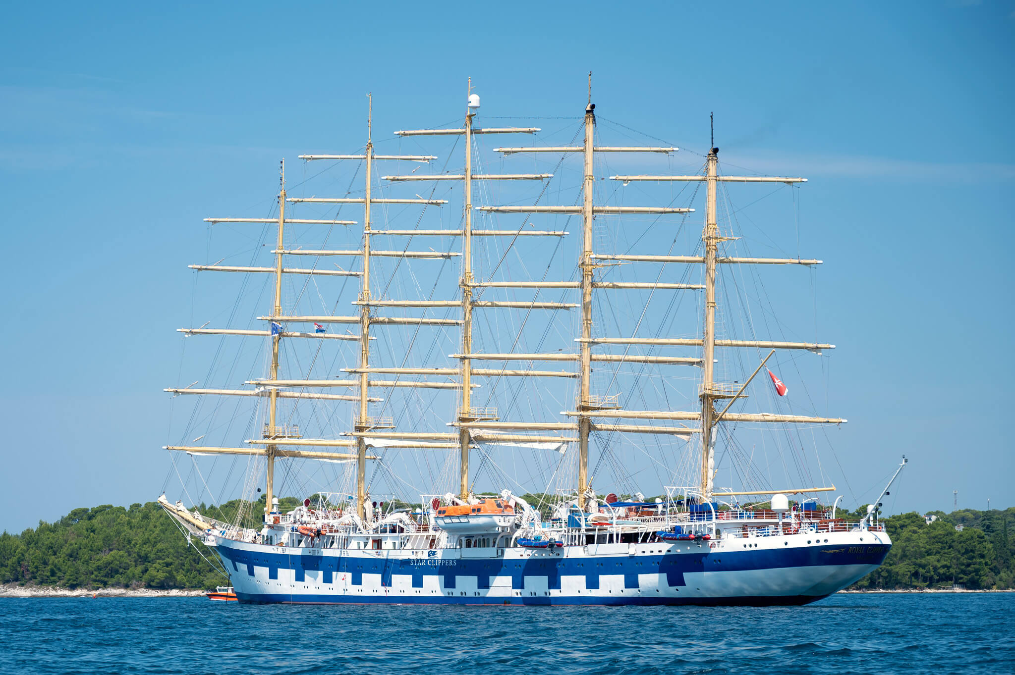 Tall-masted Royal Clipper sailing ship with multiple horizontal yards on a blue sea, hull painted white and blue, coastline in background.