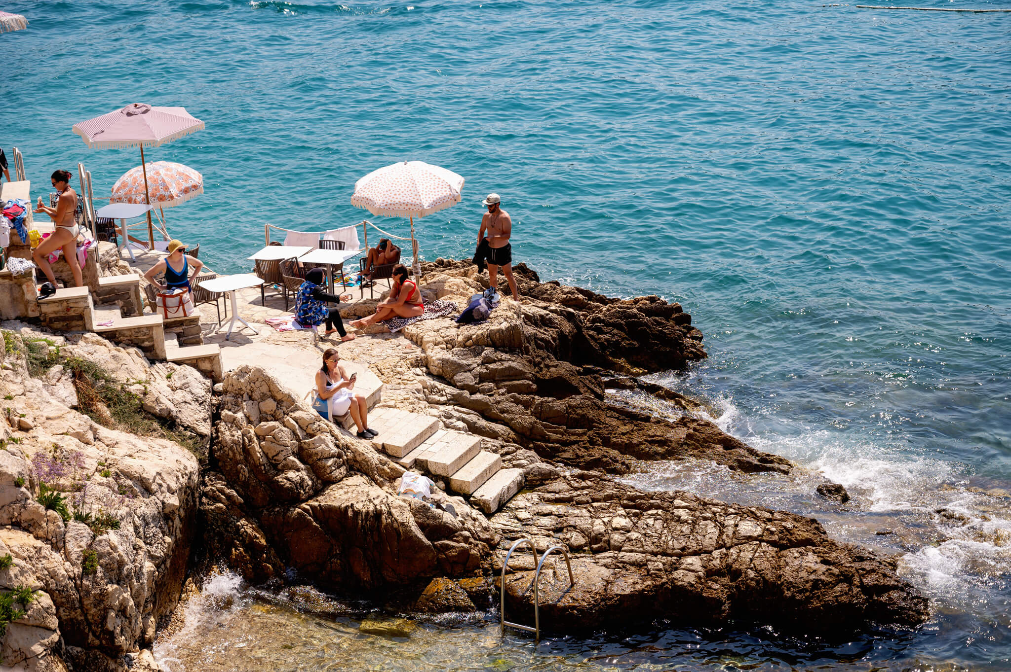 Star Clippers Royal Clipper Day 6 Rovinj Croatia People lounging on rocky coastline beside a small seating area under patterned umbrellas overlooking the blue sea.