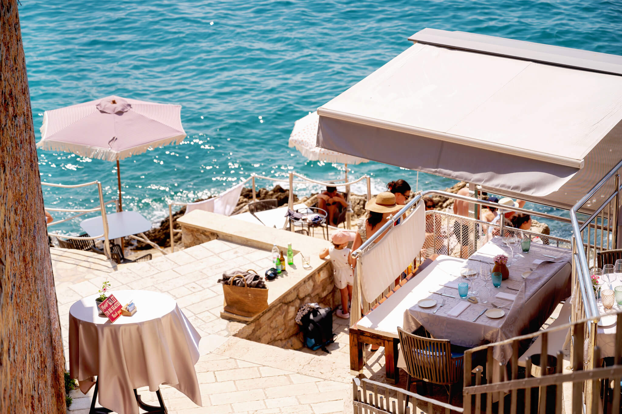 Star Clippers Royal Clipper Day 6 Rovinj Croatia Outdoor seaside restaurant terrace with white-tablecloth tables, pink and white umbrellas, and turquoise sea in the background, sunlit and relaxed.