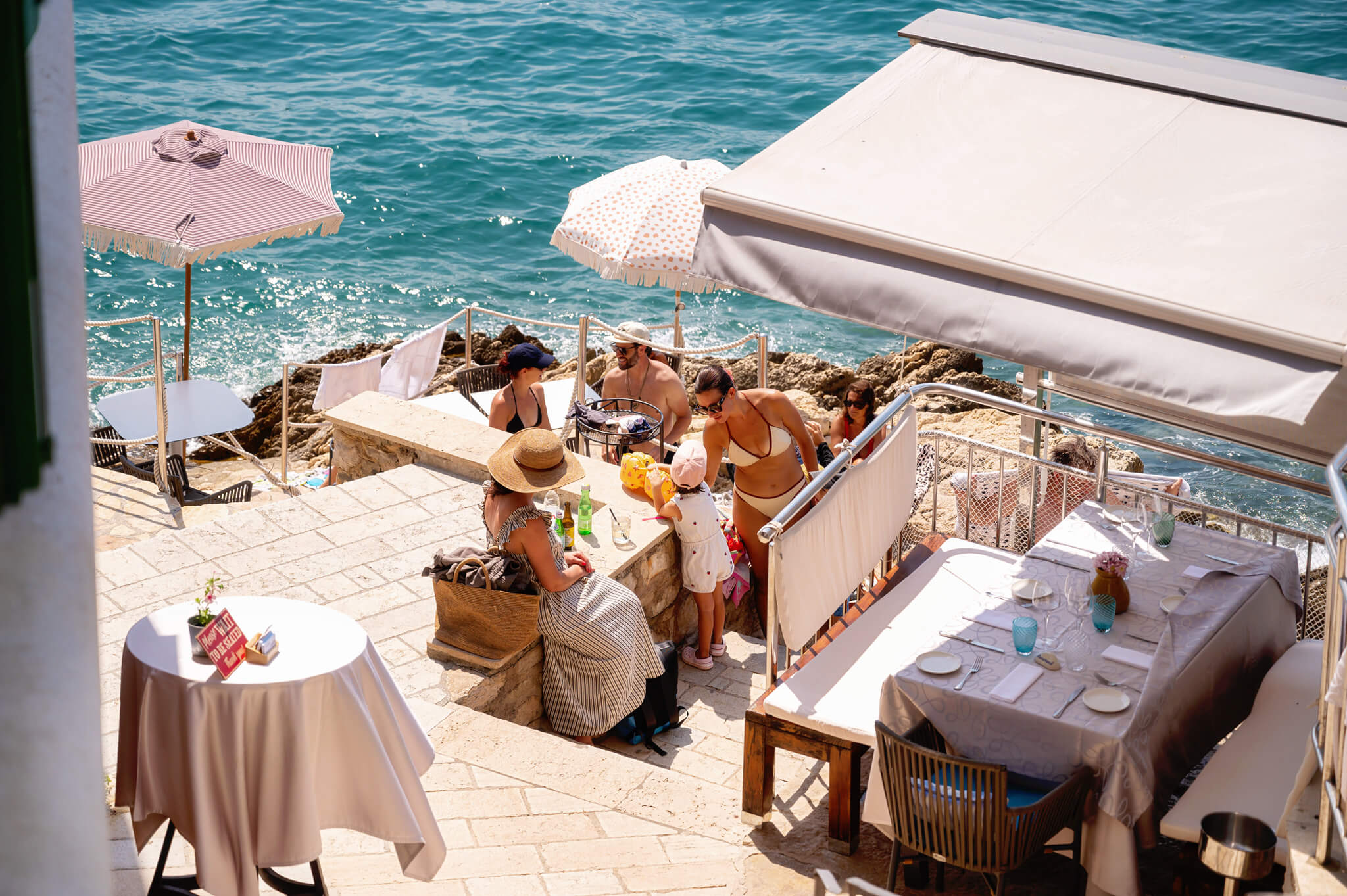 Star Clippers Royal Clipper Day 6 Rovinj Croatia 20 • Our Little Lifestyle Outdoor seaside restaurant on a stone terrace with white-tablecloth tables, striped umbrellas, and guests in swimwear by the blue water. Star Clippers Royal Clipper Day 6 Rovinj Croatia