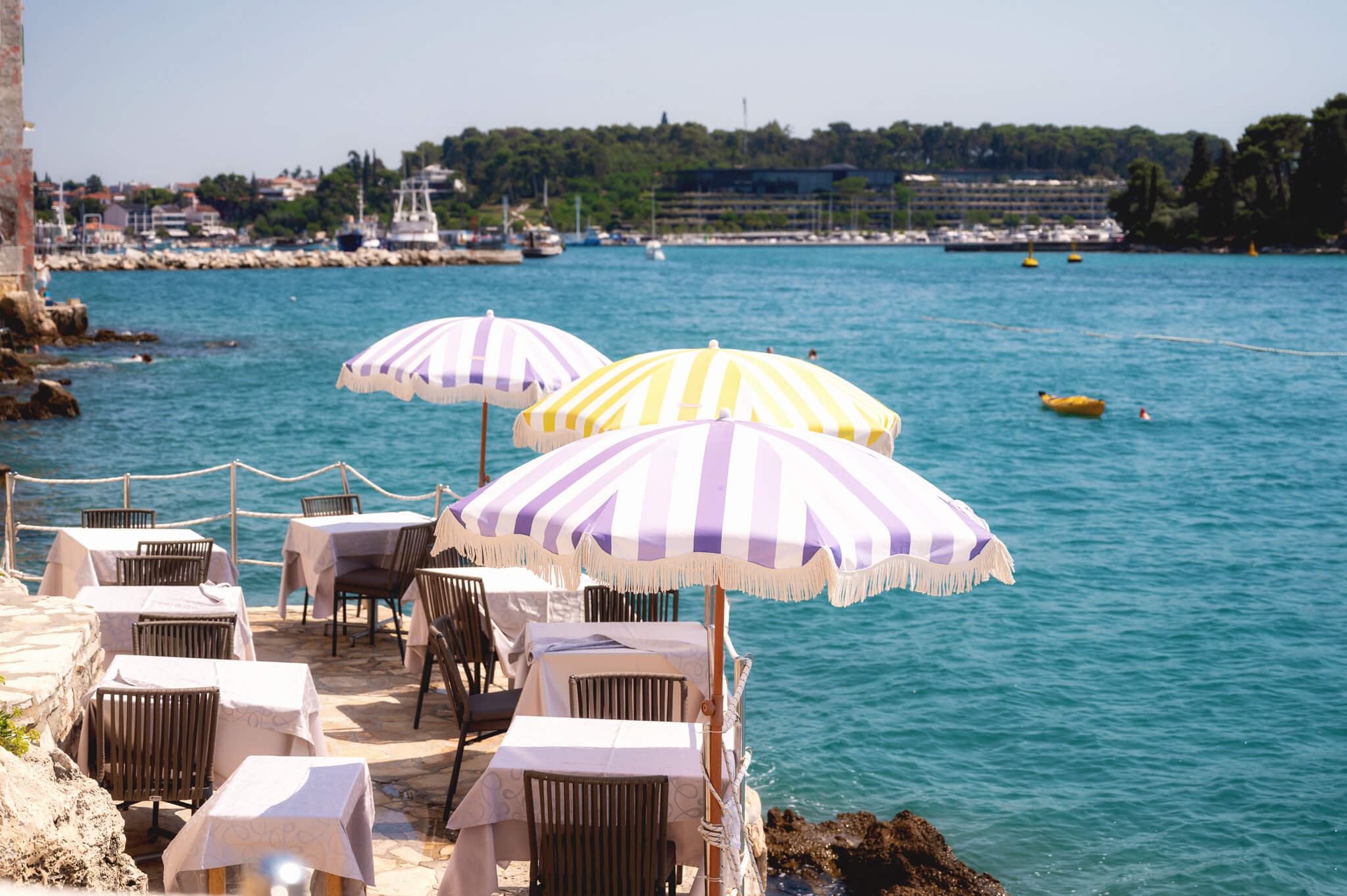 Seaside restaurant setup with white tables and purple-striped umbrellas overlooking a turquoise bay. Star Clippers Royal Clipper Day 6 Rovinj Croatia