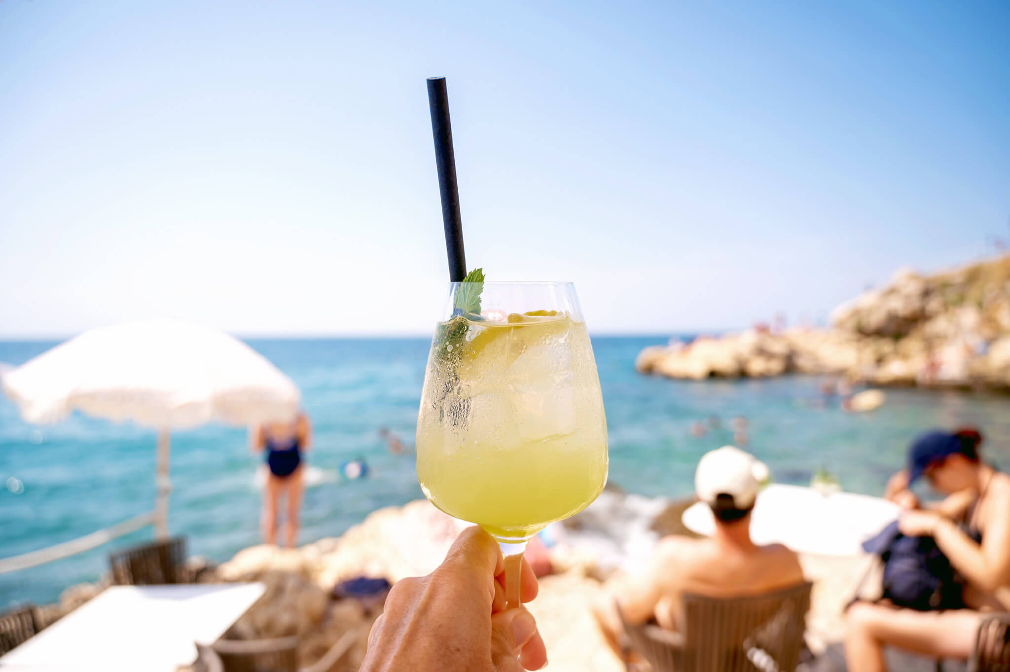 Hand holding a citrus cocktail with a black straw at a sunny beach bar, sea and rocks in the background. Star Clippers Royal Clipper Day 6 Rovinj Croatia