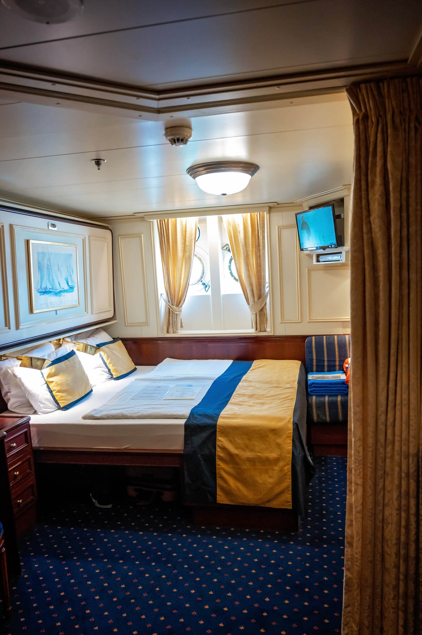 Hotel ship cabin with a large bed dressed in yellow, navy, and white linens, gold curtains, and a wall-mounted TV.