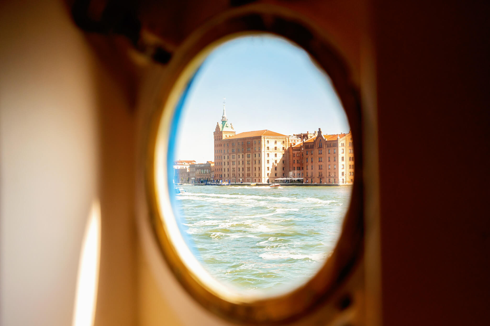 View of a waterfront city seen through a circular porthole, with brick buildings along the river and a green-roofed tower in the background on a sunny day