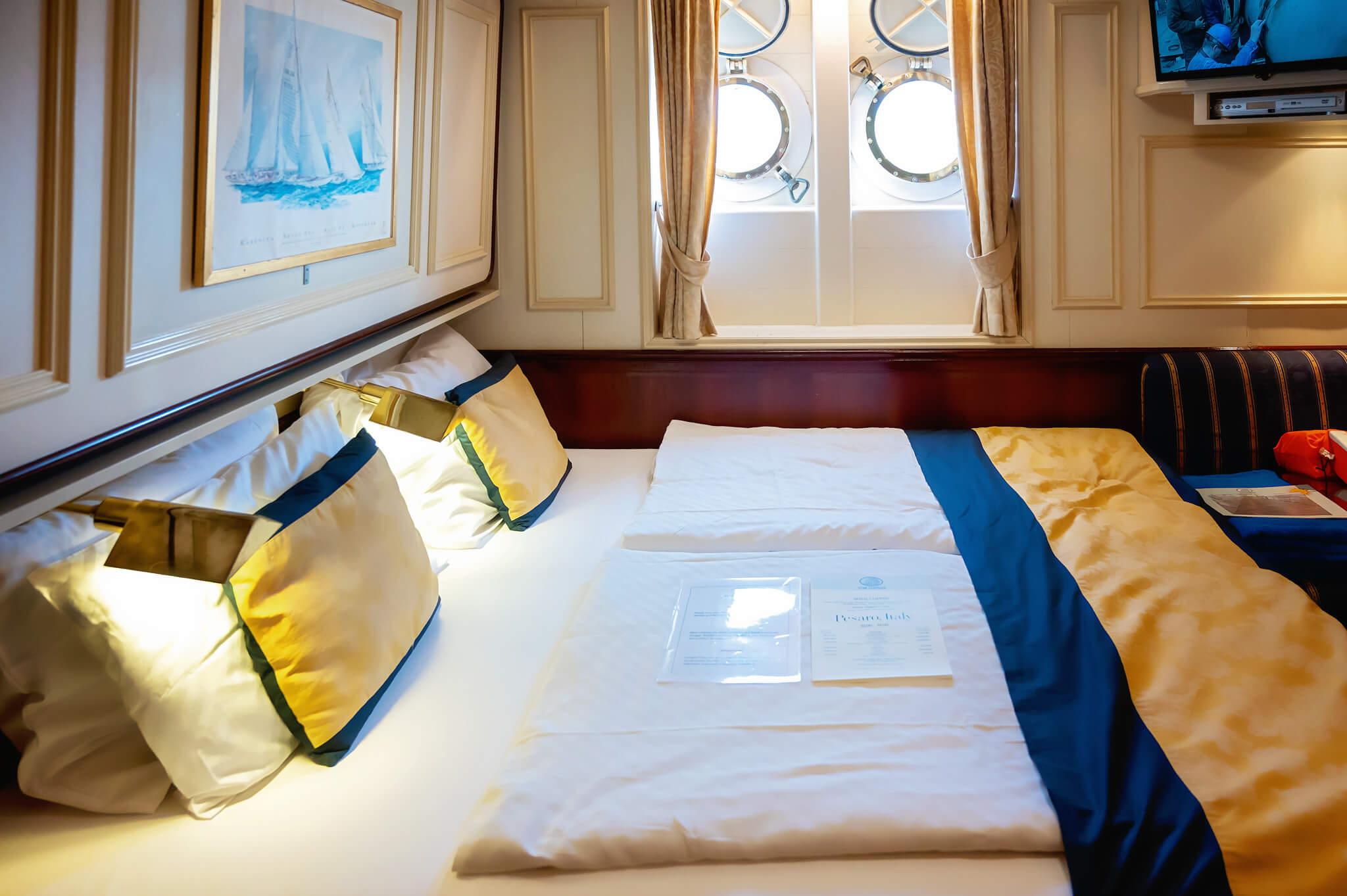 A cozy ship cabin with a bed, cream bedding and yellow-green-blue pillows, wood trim, and round porthole windows at the far wall with beige curtains.