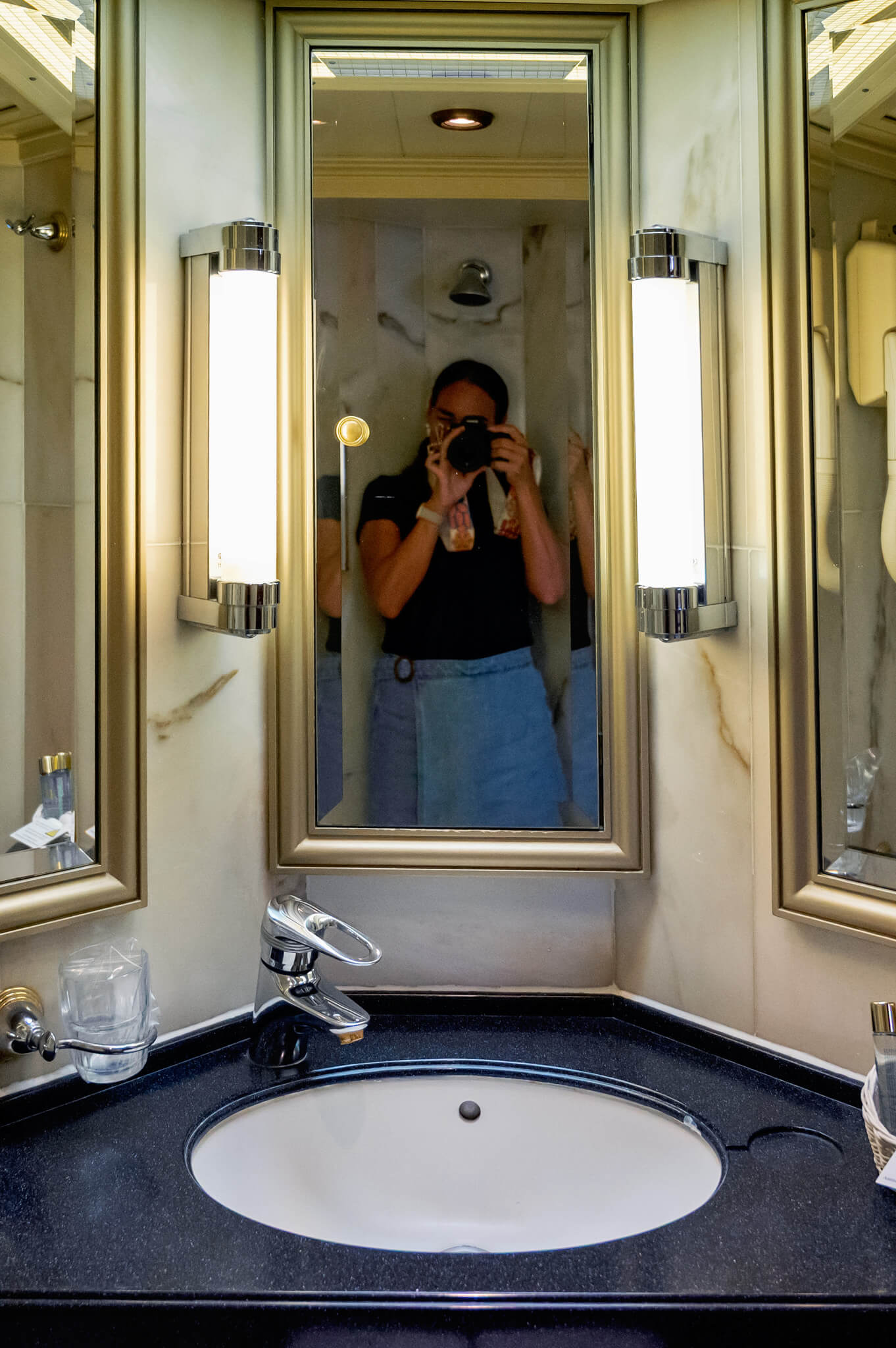 Person taking a mirror selfie in a marble bathroom; central mirror flanked by lit vanity mirrors on both sides