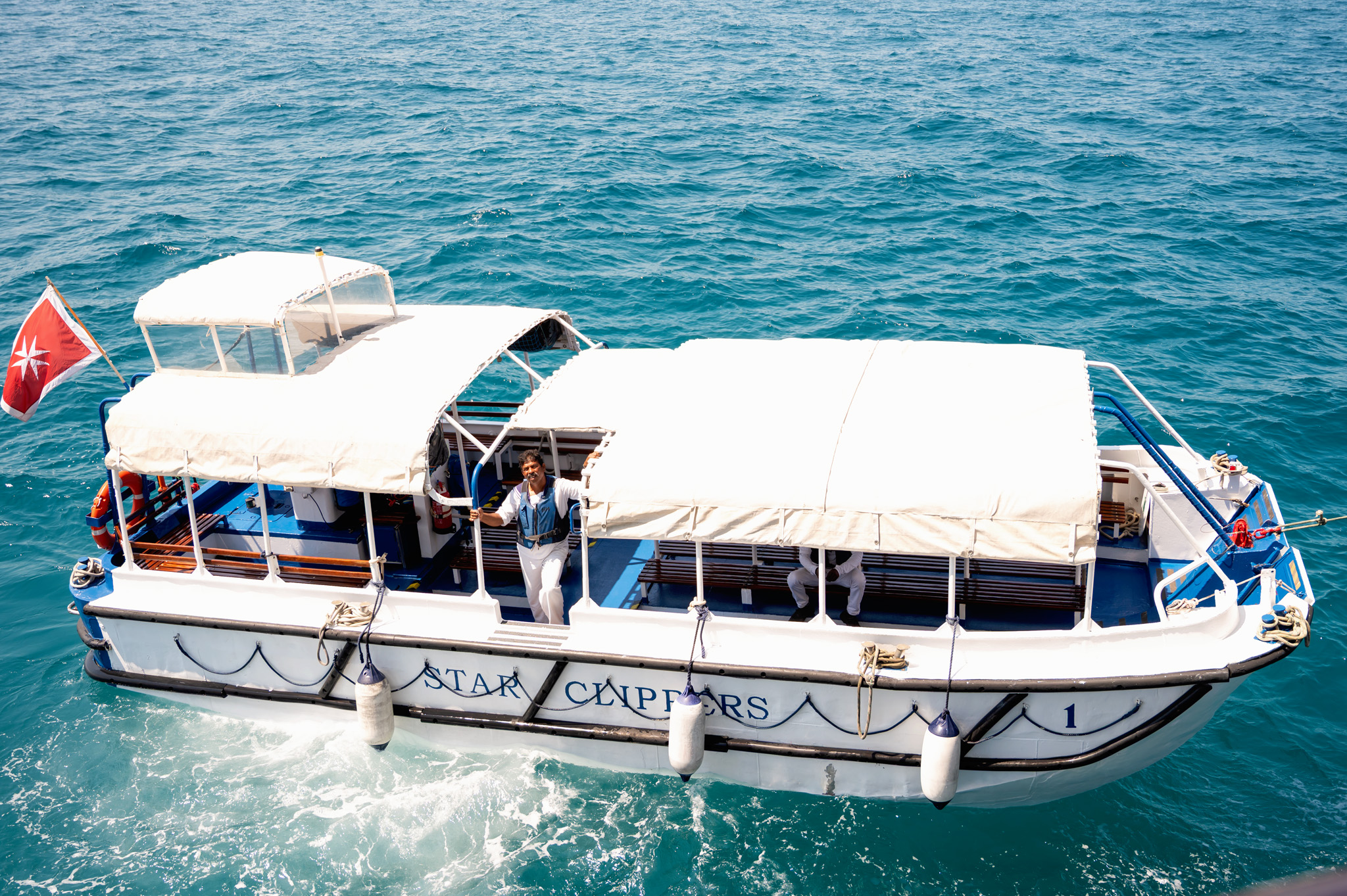 Aerial view of a white cruise boat on turquoise ocean with a white canopy and passengers on deck under the canopy, flag at the stern