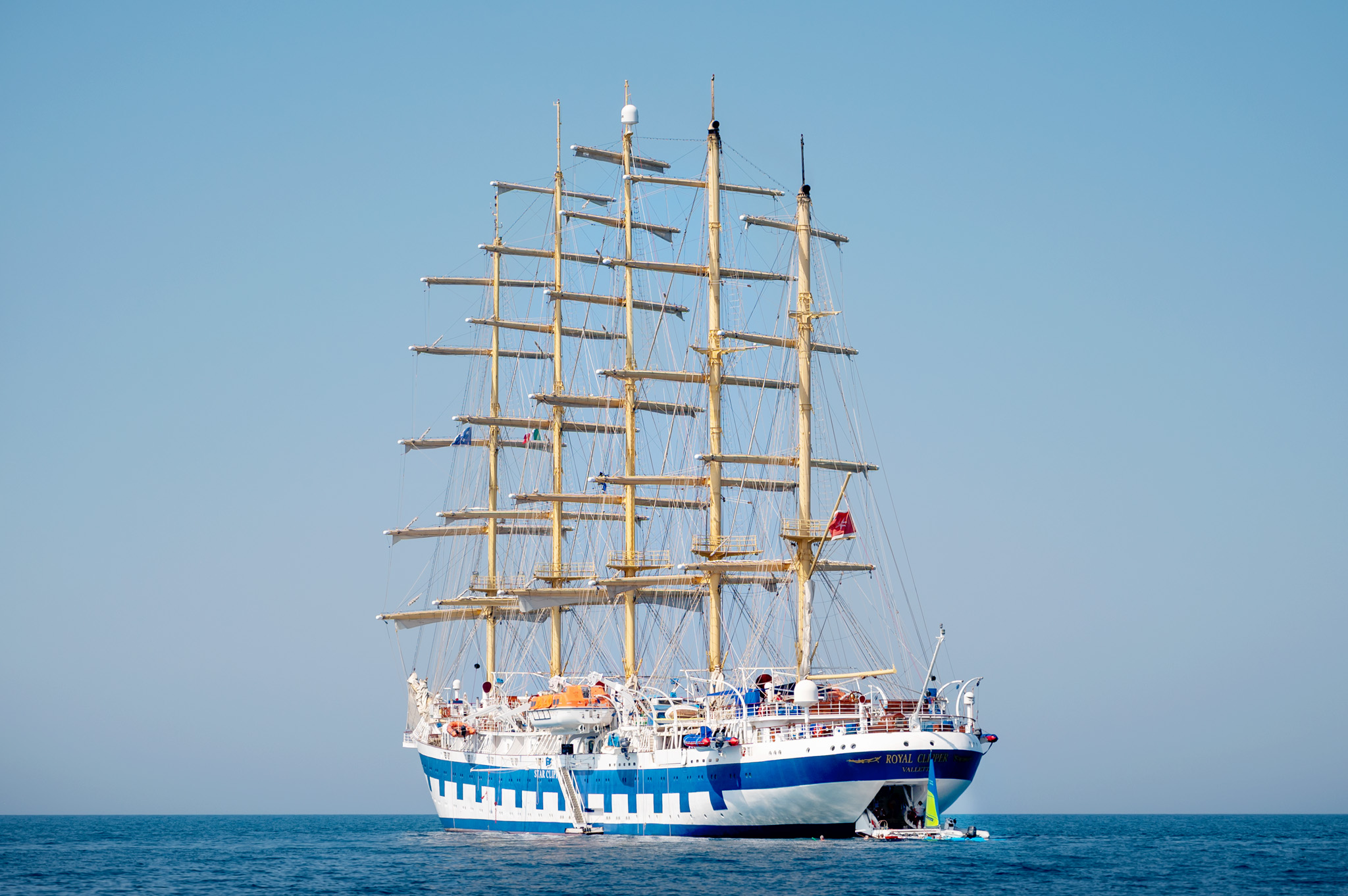 Four-masted sailing cruise ship with a blue and white hull on calm blue sea, clear sky above
