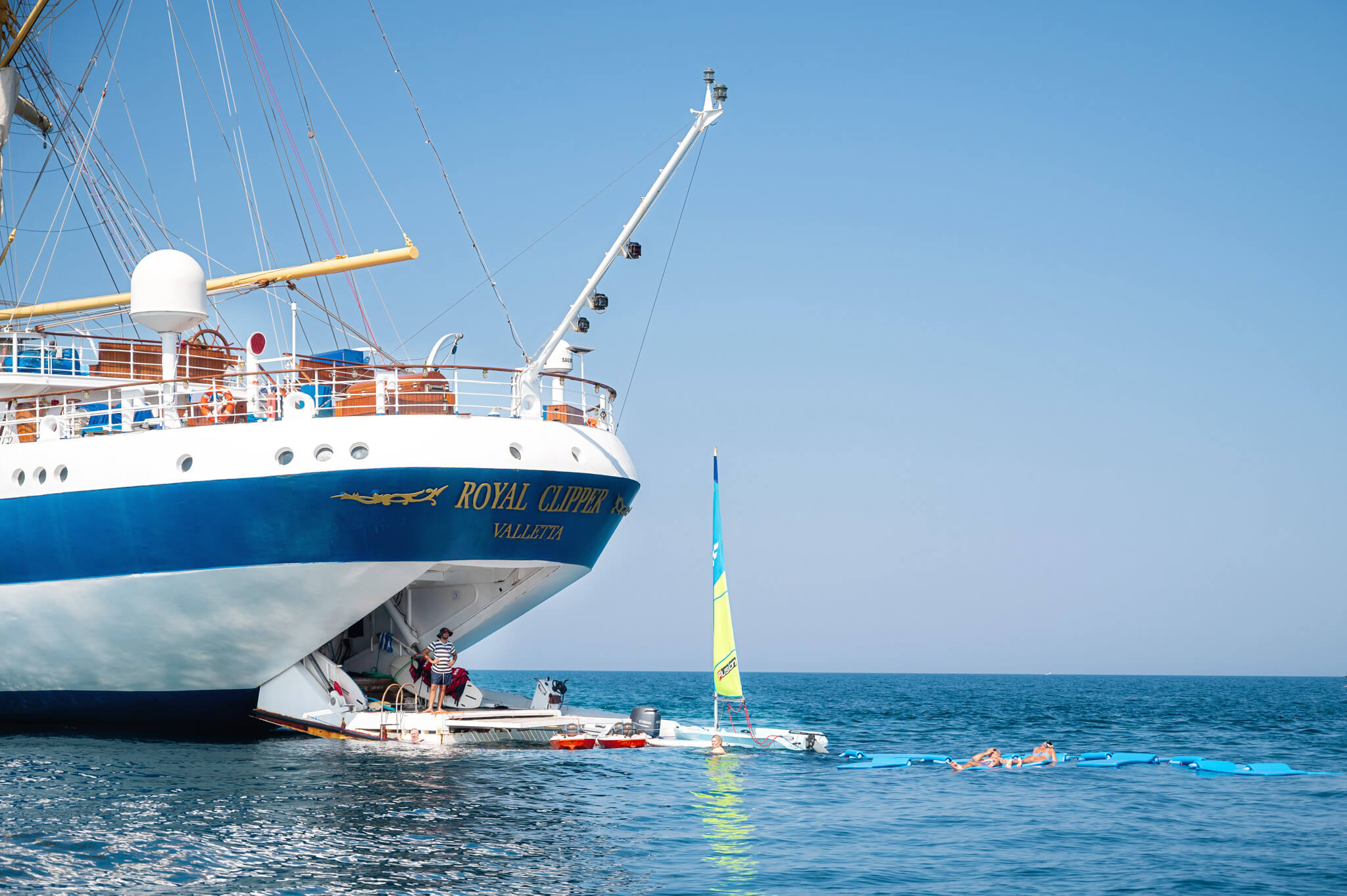 Bow of a white-and-blue cruise ship named Royal Clipper with passengers on deck, calm sea, and a clear blue sky