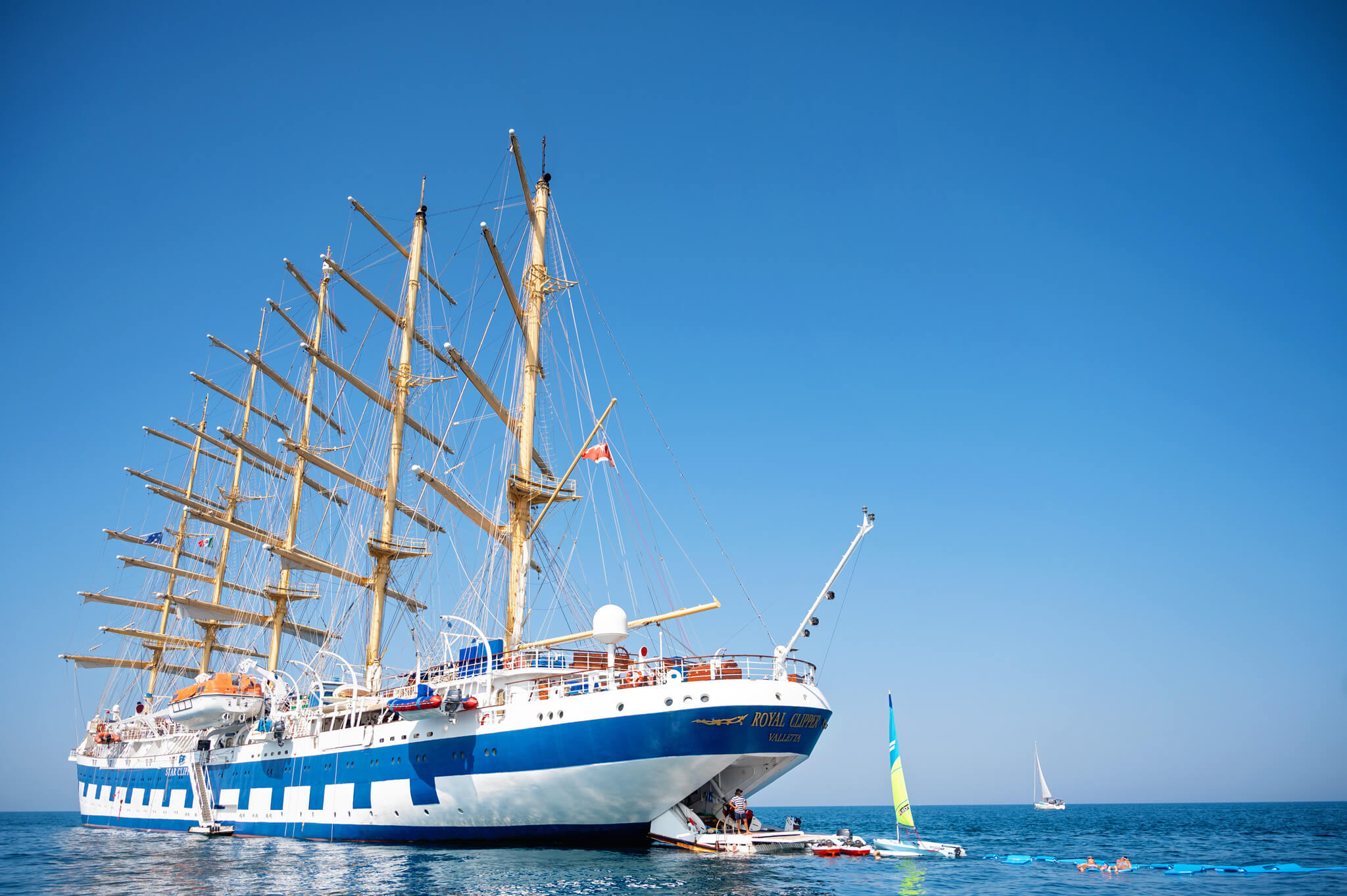 Royal Clipper cruise ship with tall masts and rigging docked beside clear blue water on a sunny day