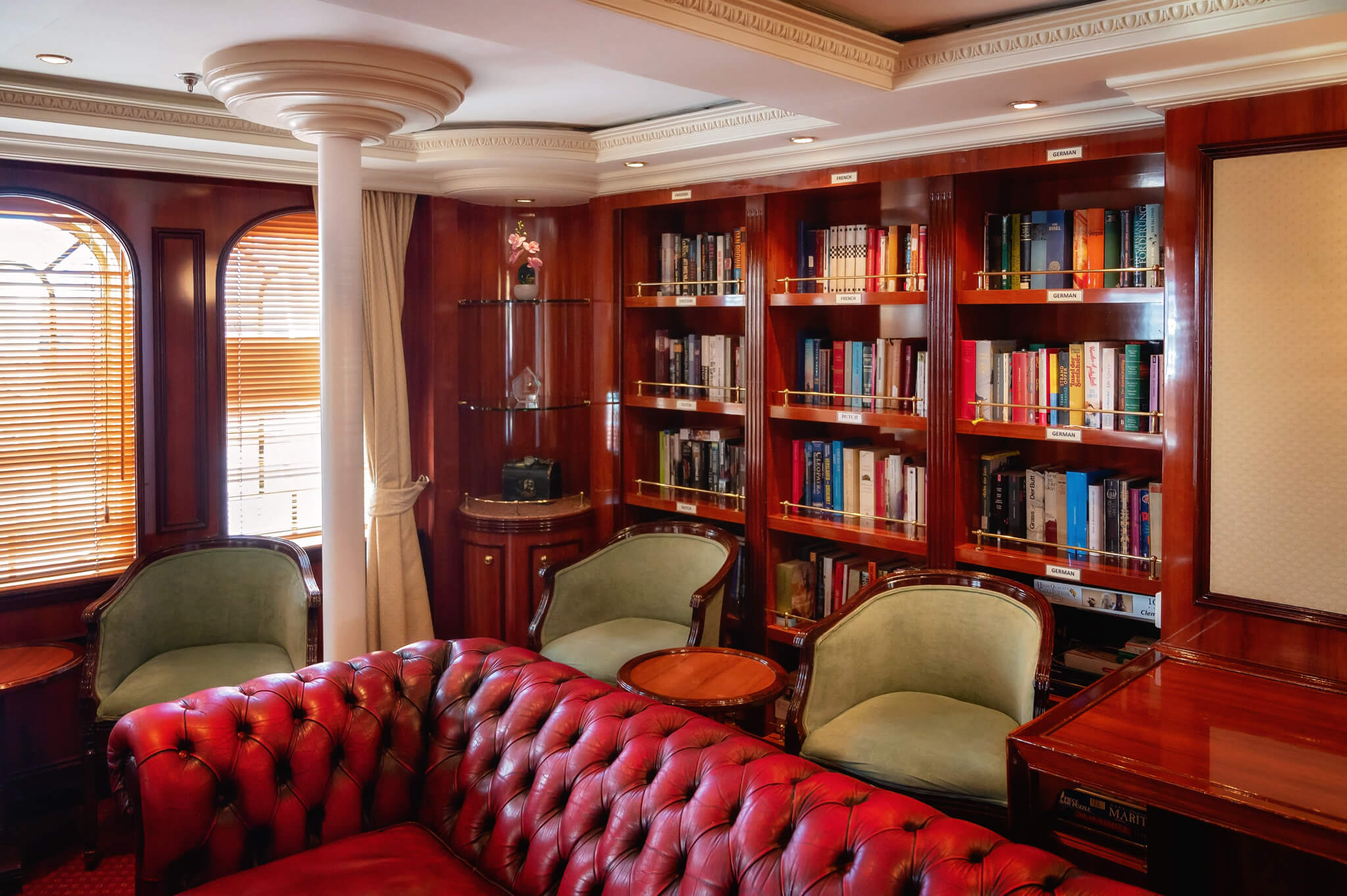 Luxurious library lounge with a red tufted leather sofa, green upholstered chairs, and tall dark wood bookshelves.