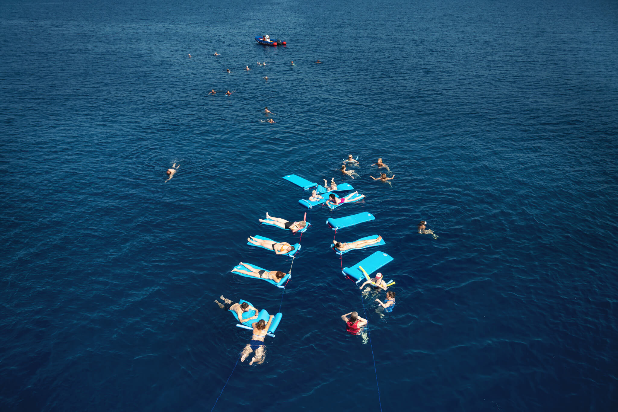 Overhead view of people on bright blue float mats lounging in deep blue ocean, with a small motorboat nearby.